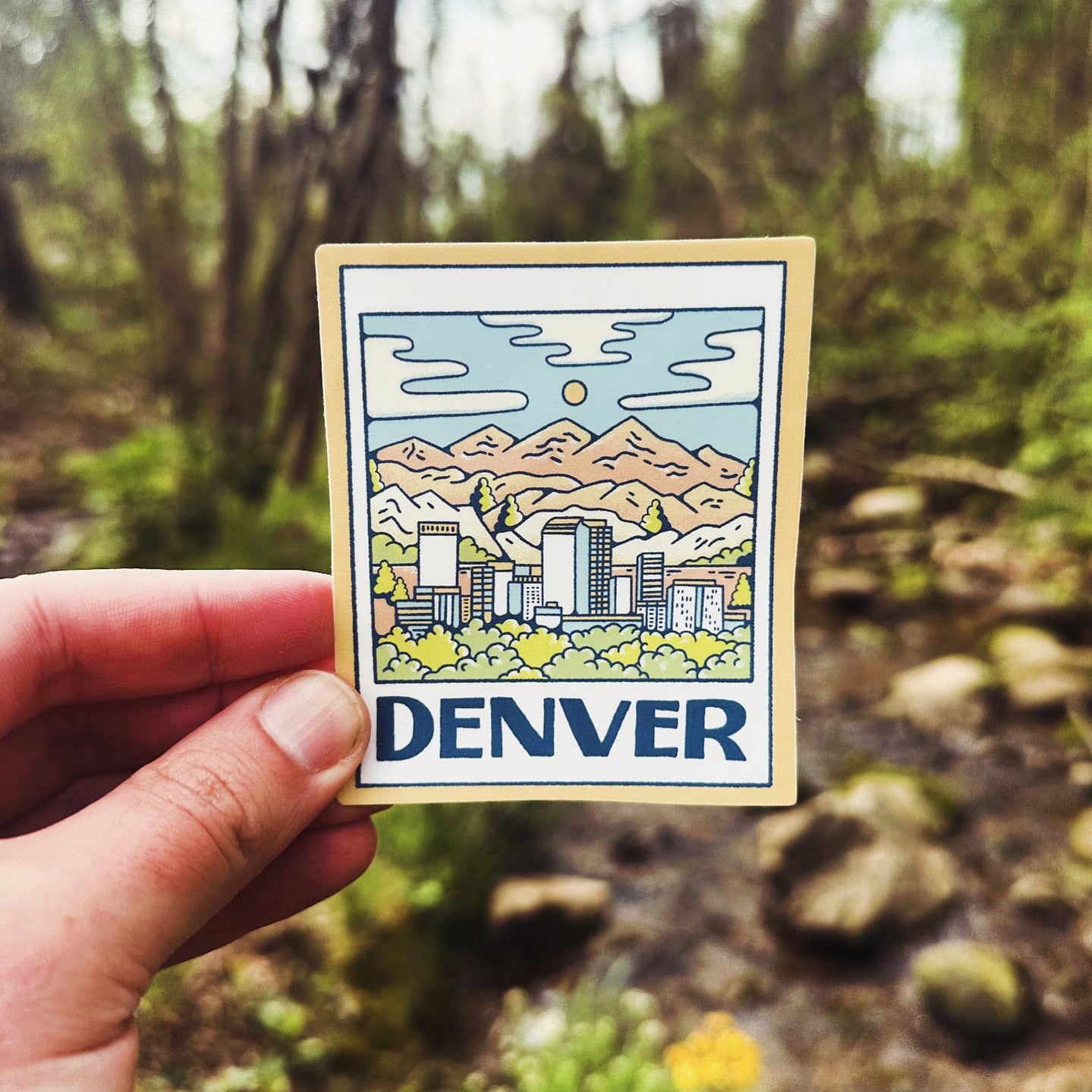 Hand holding a sticker with a mountain and cityscape design labeled 'Denver' against a natural background.