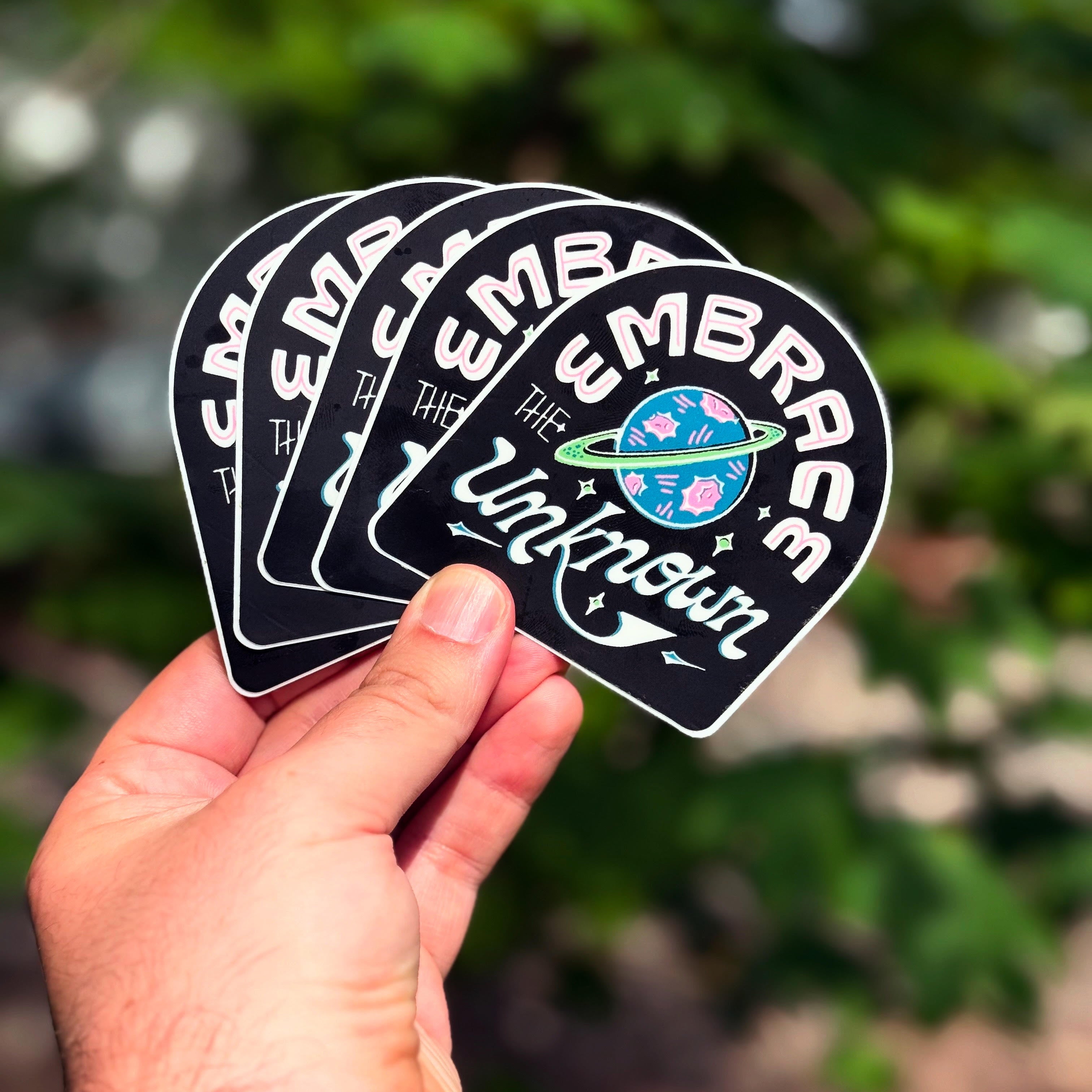 Hand holding four 'Embrace the Unknown' stickers with a blurred green background