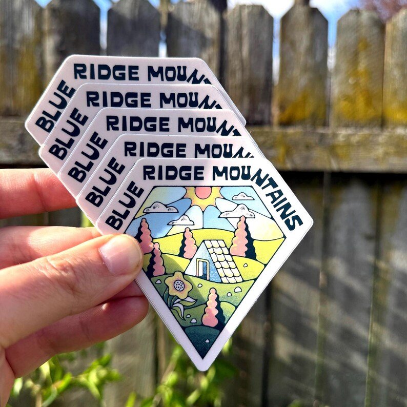 Hand holding a set of Blue Ridge Mountains-themed stickers against a wooden fence.