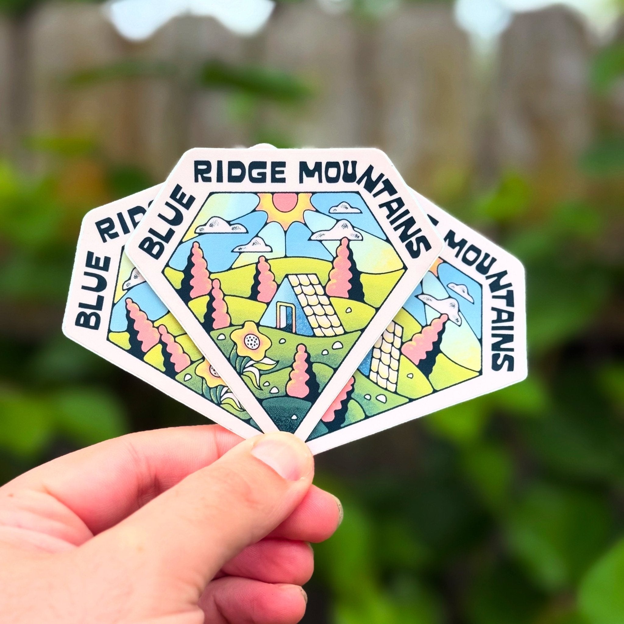 Hexagonal sticker with 'Blue Ridge Mountains' text and nature illustration held by a hand against a blurred green background.