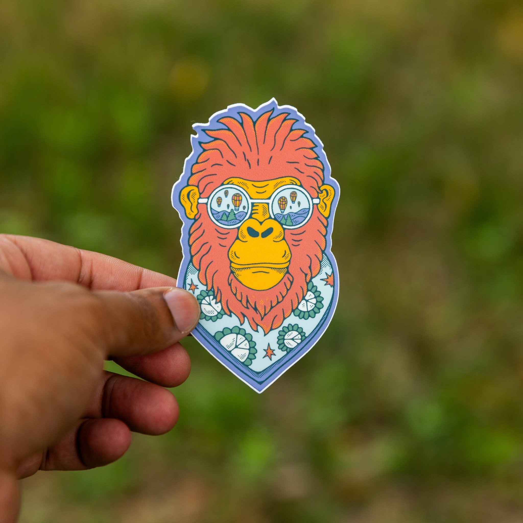 Close-up of a hand holding a vinyl sticker of a red-orange Bigfoot wearing balloon sunglasses and a patterned scarf, with a blurred green background.