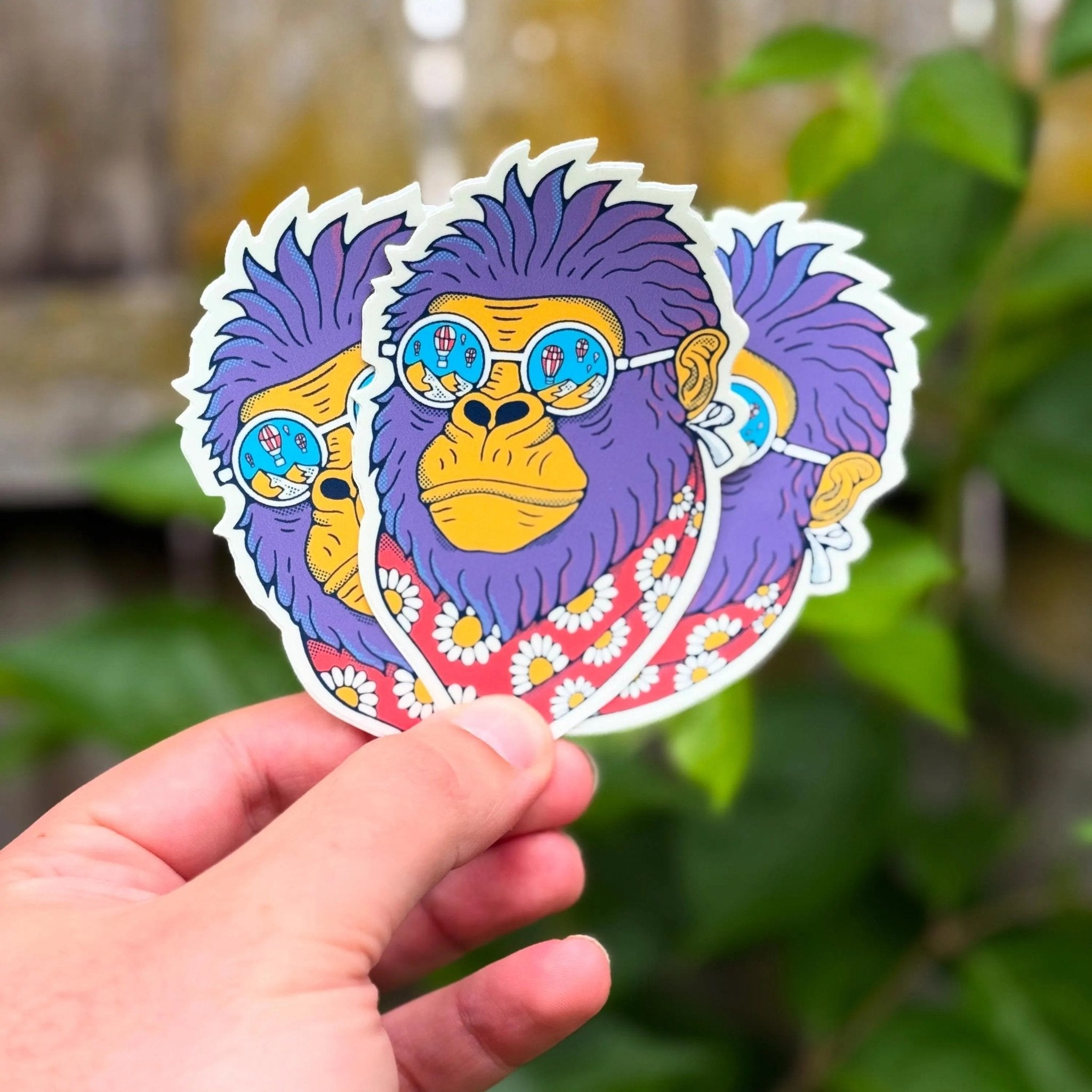 Colorful sticker of a monkey with sunglasses held by a hand against a blurred green background