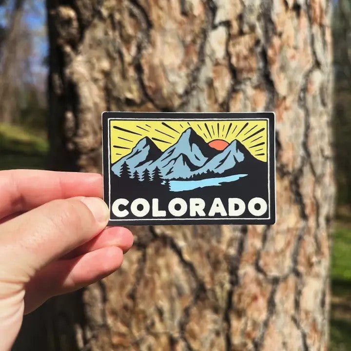 A hand holding a vinyl sticker with the image of mountains and the text 'COLORADO' on it, with a background of a tree trunk.