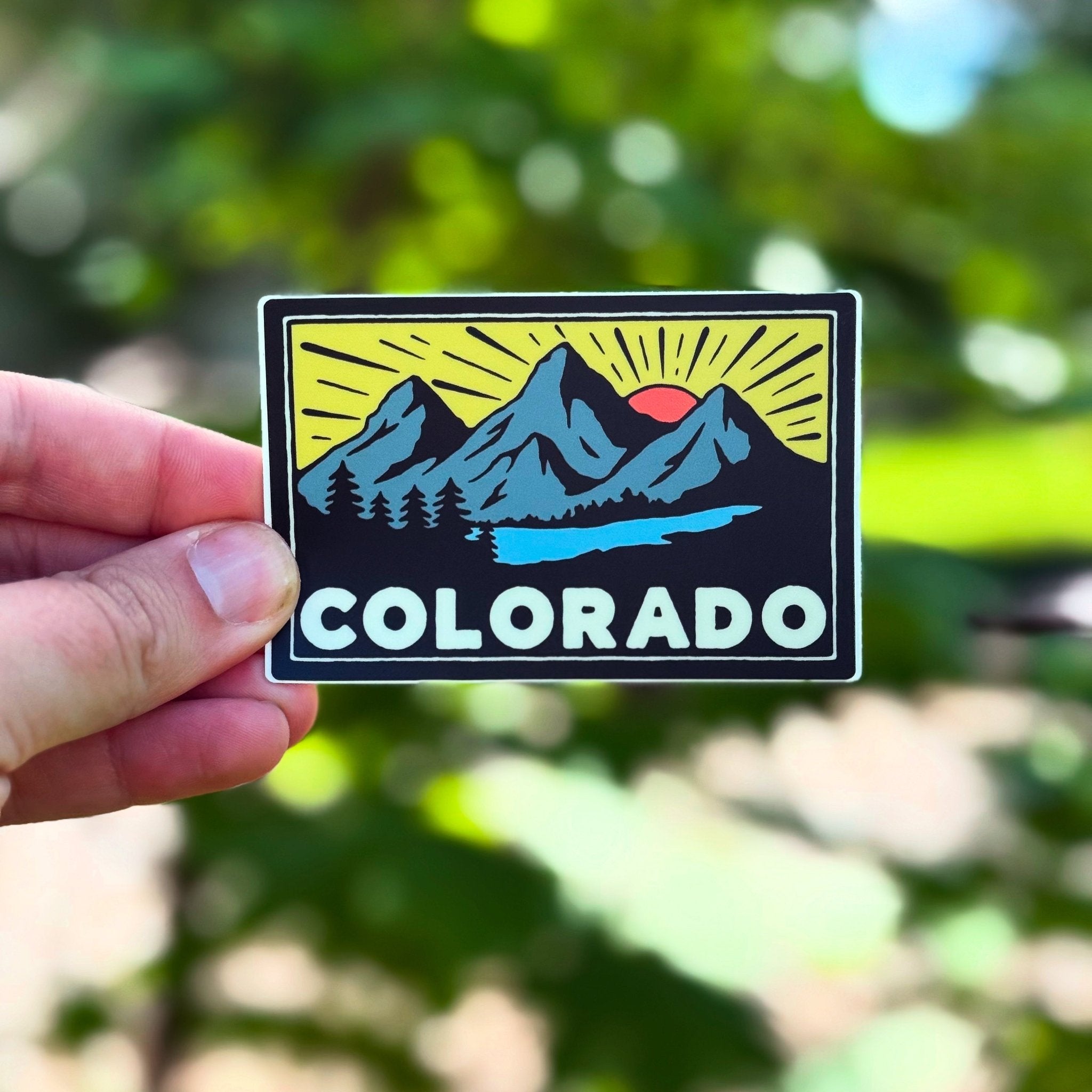 Hand holding a 'Colorado' sticker with mountain design against a blurred green background
