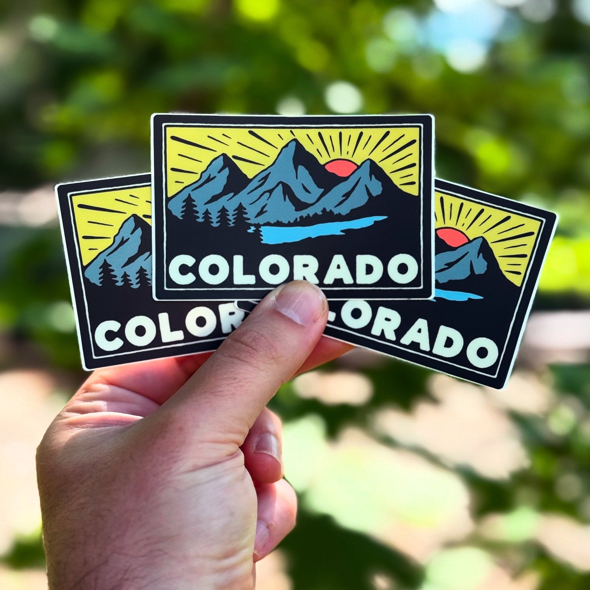 Hand holding three 'Colorado' stickers with mountain design against a blurred green background