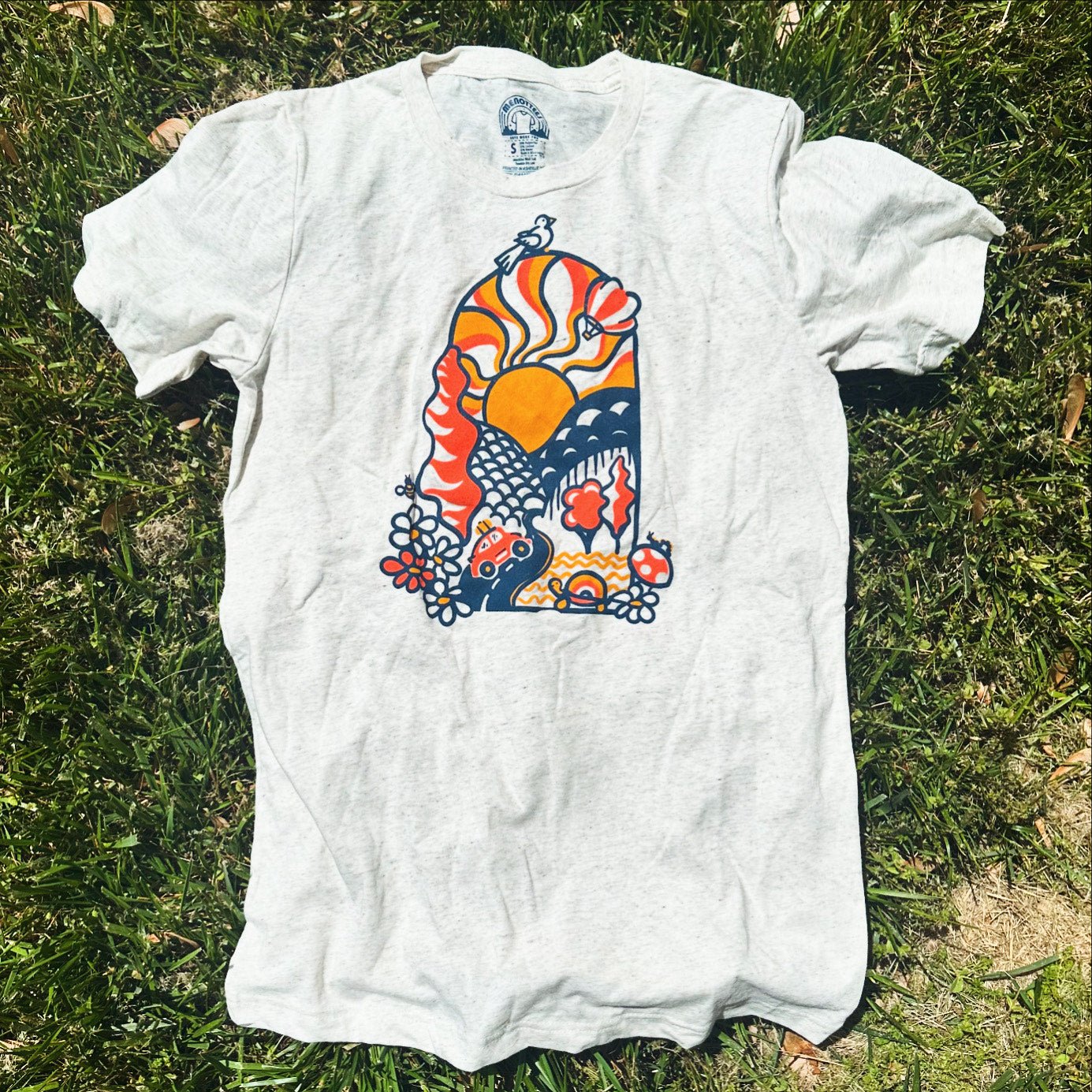 A white crew neck t-shirt with a colorful graphic design lying on the grass.
