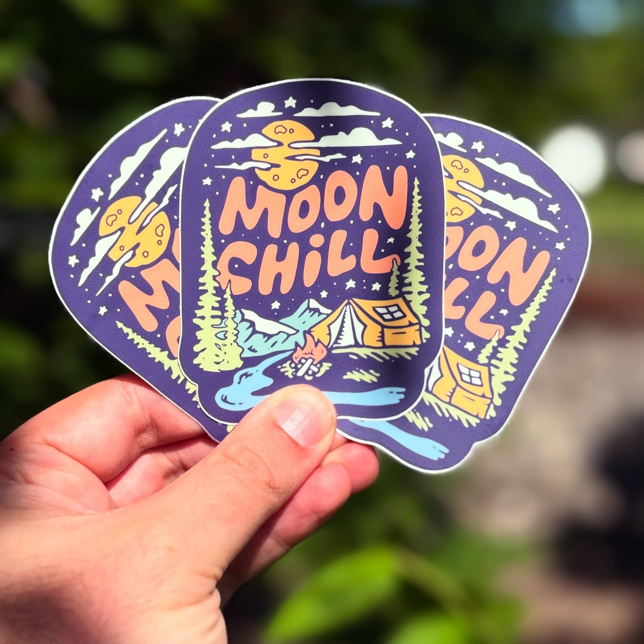Hand holding a 'Moon Chill' sticker with nature-themed design against a blurred green background