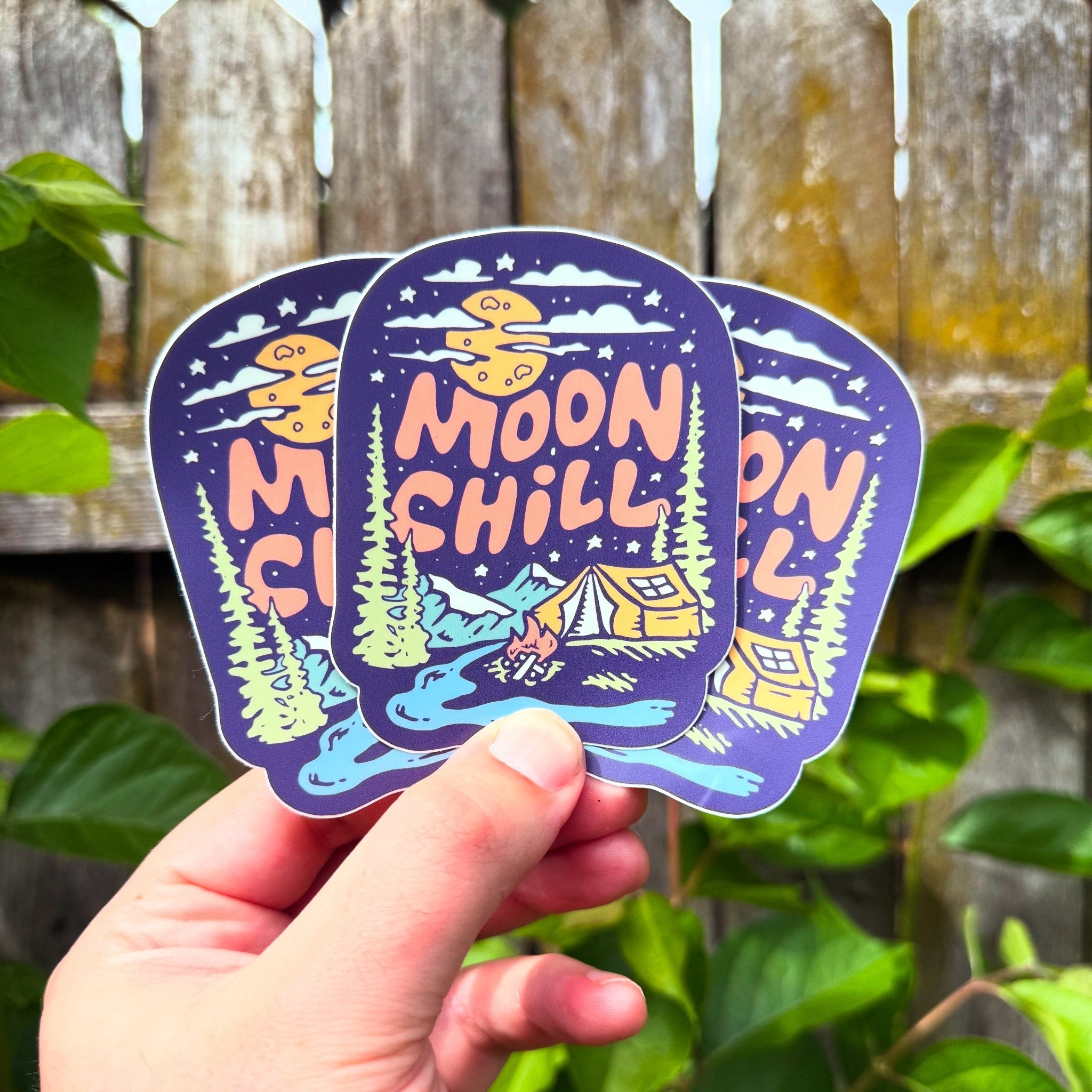 Three 'Moon Chill' stickers held by a hand with a wooden fence and greenery in the background.