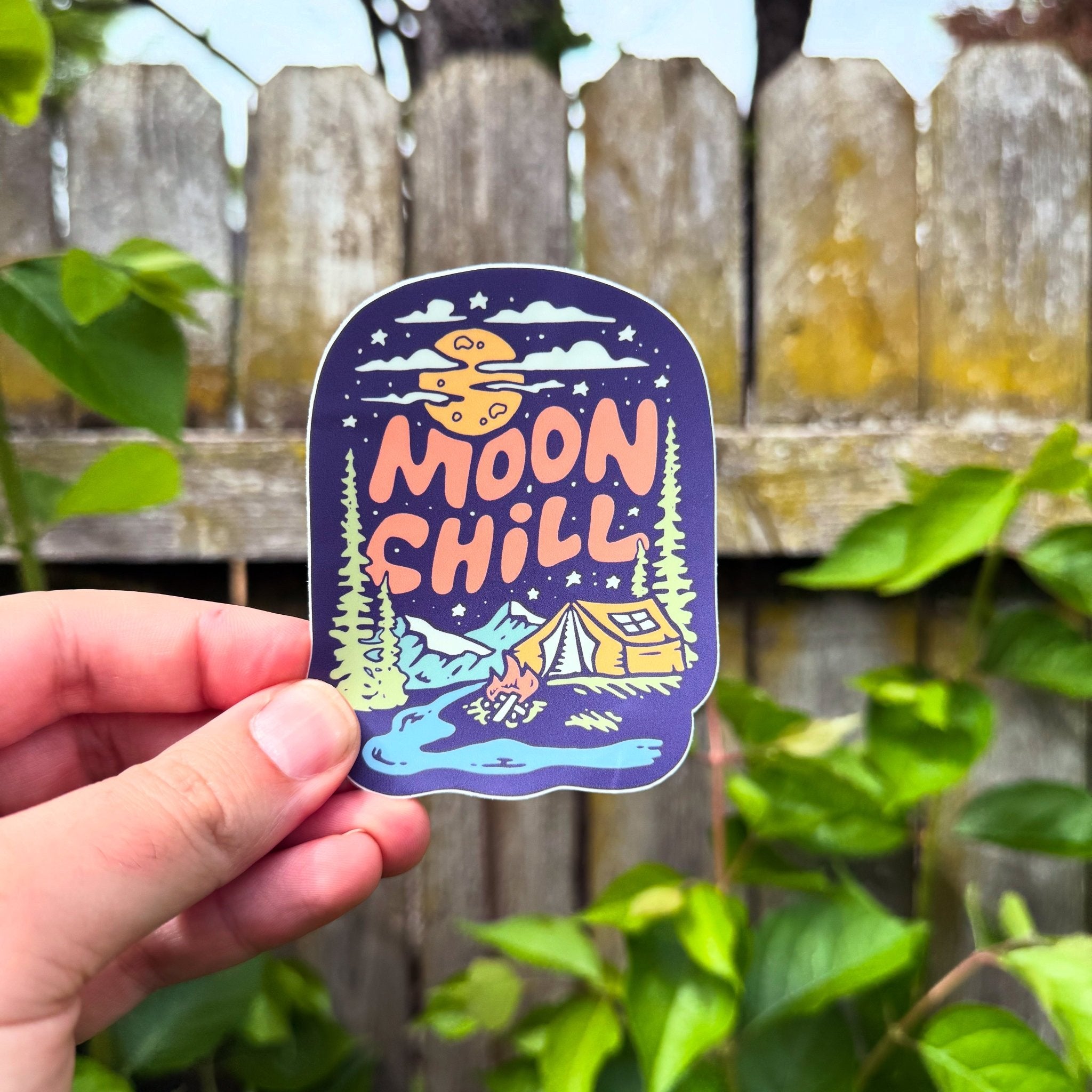 Sticker with 'Moon Chill' design held by a hand against a wooden fence and greenery background