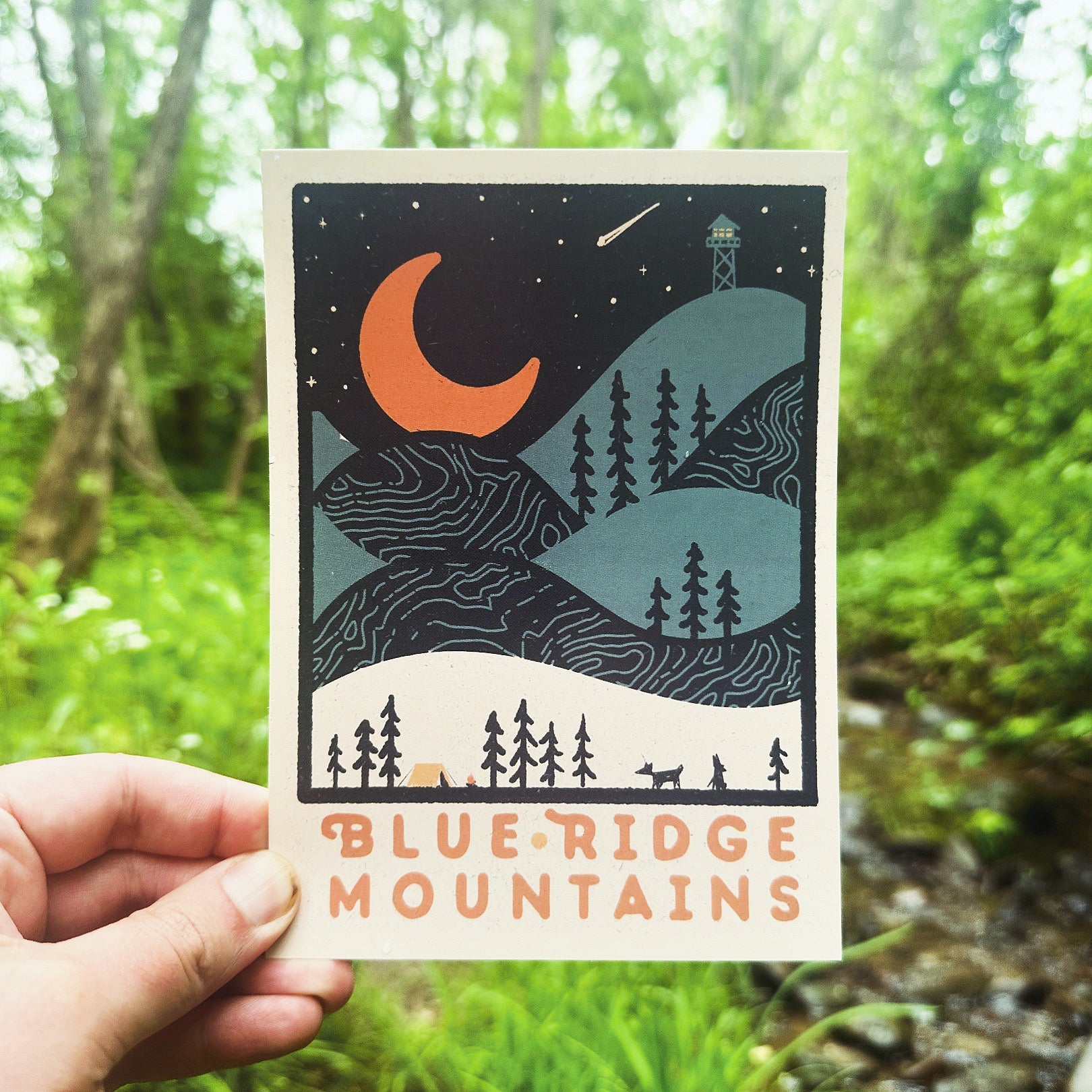 A postcard depicting a nighttime scene in the Blue Ridge Mountains with a crescent moon, trees, and a cabin.
