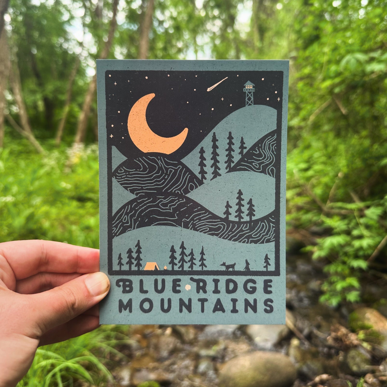 A postcard depicting a night view of the Blue Ridge Mountains with a crescent moon, trees, and a cabin, held in a hand with a blurred background.