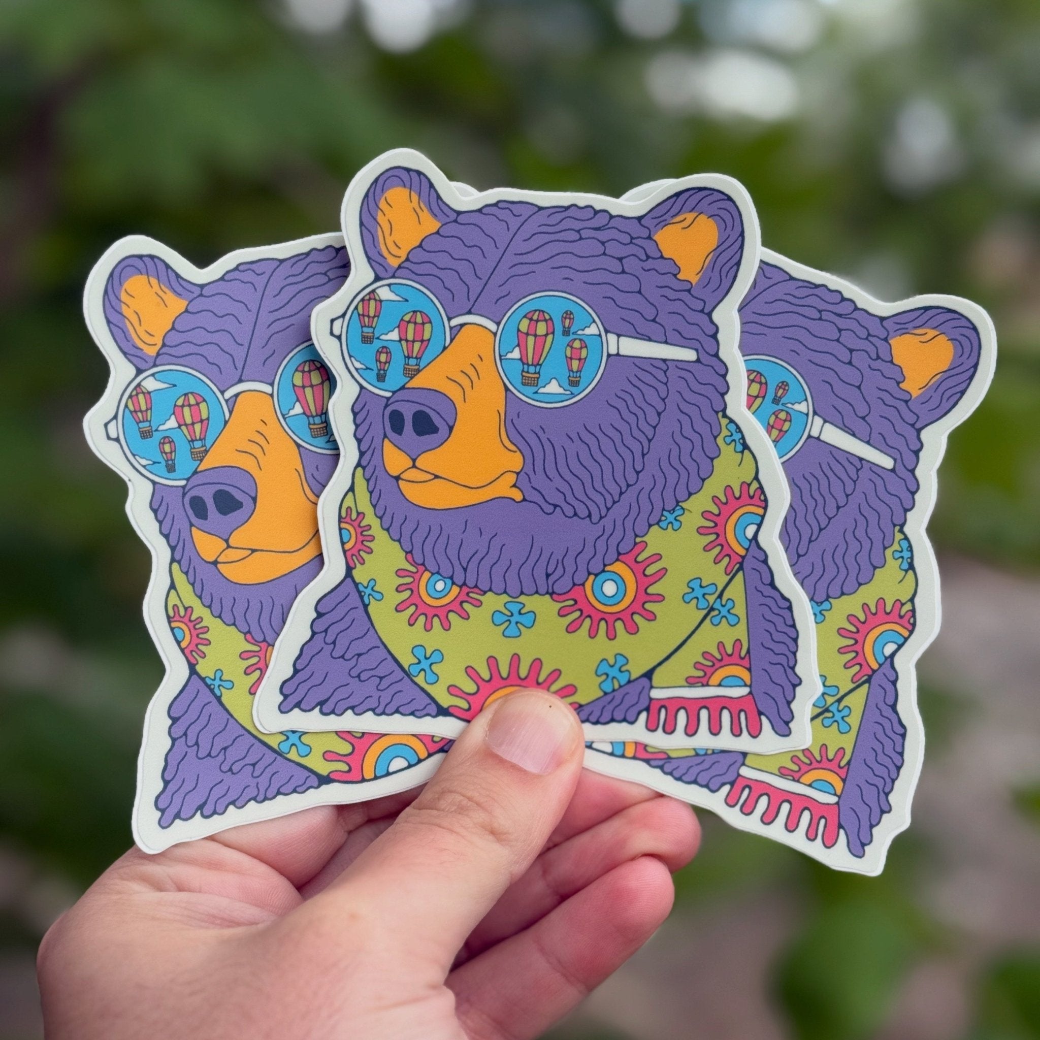Colorful bear sticker with sunglasses held by a hand against a blurred green background