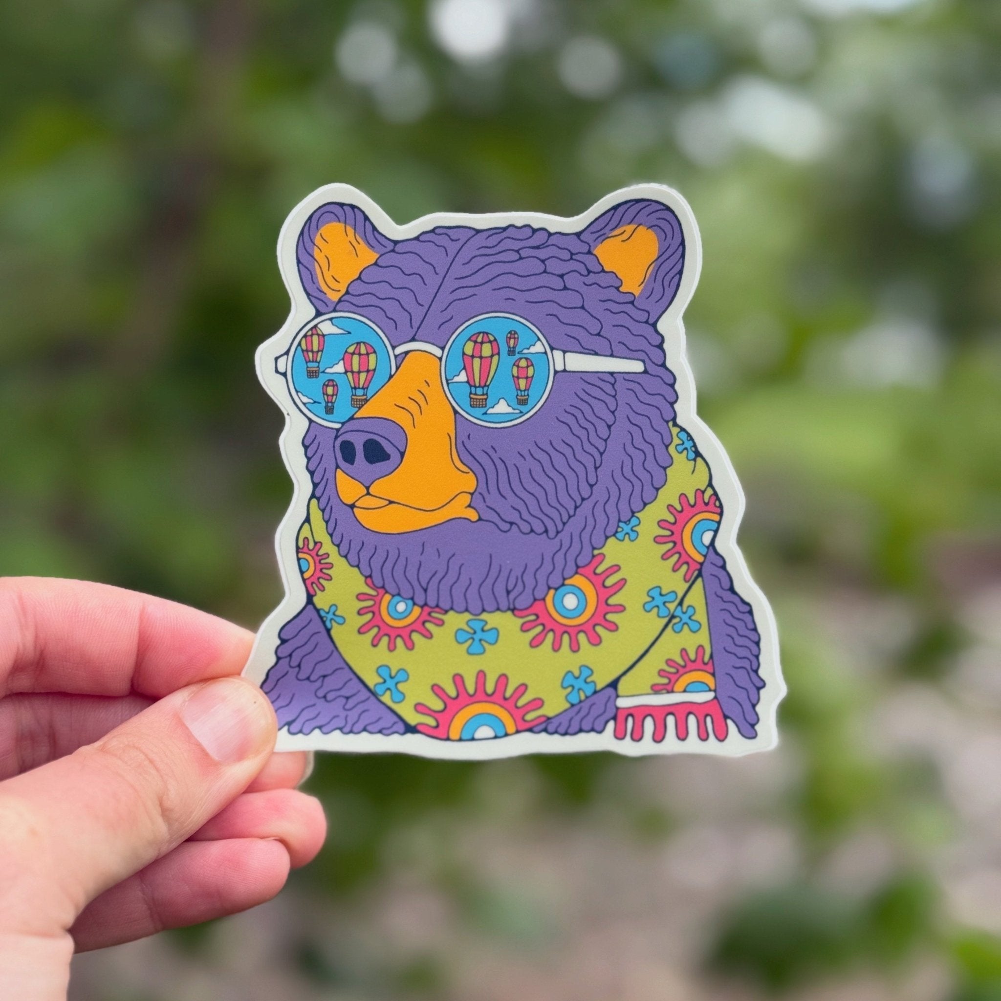 Colorful bear sticker with sunglasses held by a hand against a blurred green background