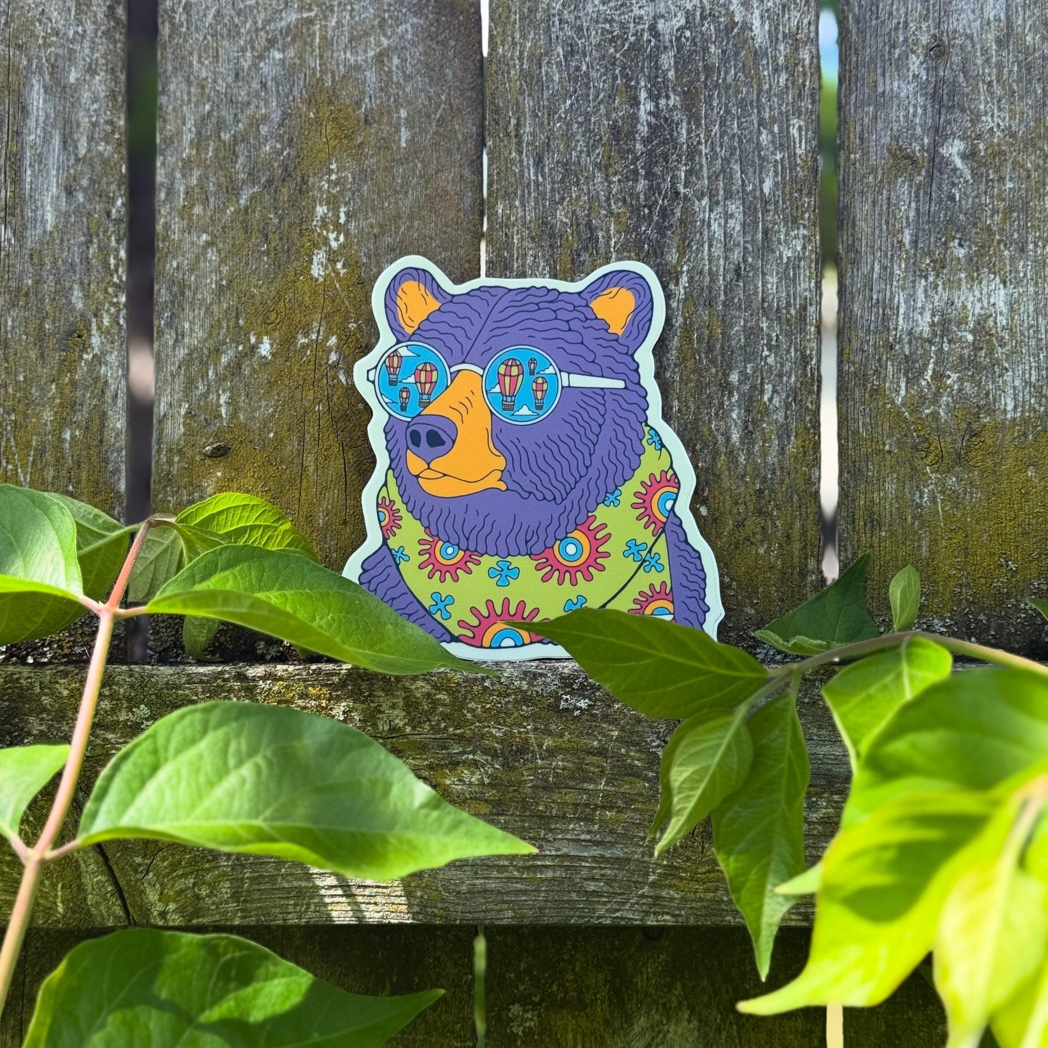 Colorful bear sticker with sunglasses on a wooden surface with green leaves.