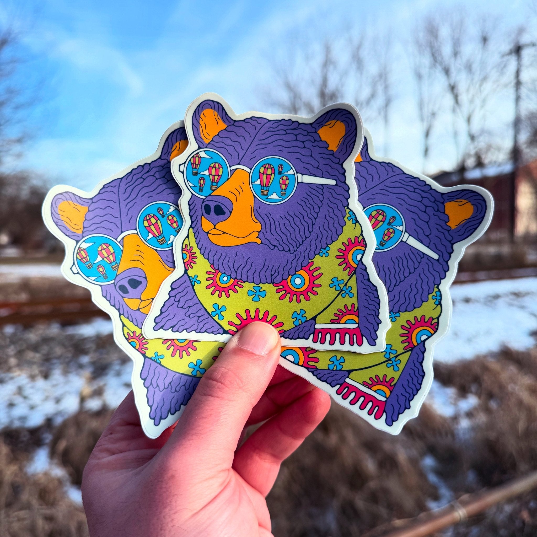 A hand holding three colorful vinyl stickers with a bear design against an outdoor background.