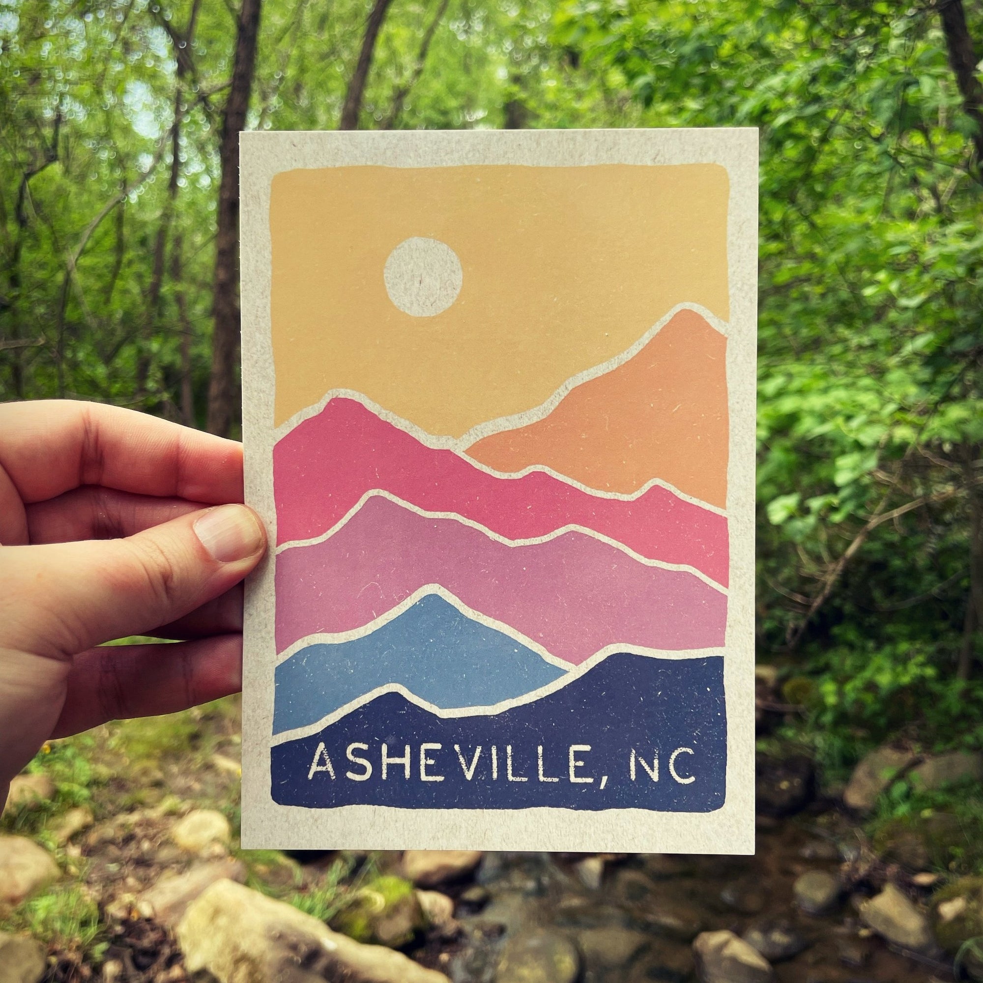 A postcard depicting a stylized mountain range in colors from pink to blue, with the text 'Asheville, NC' at the bottom, held in a hand with a blurred natural background.