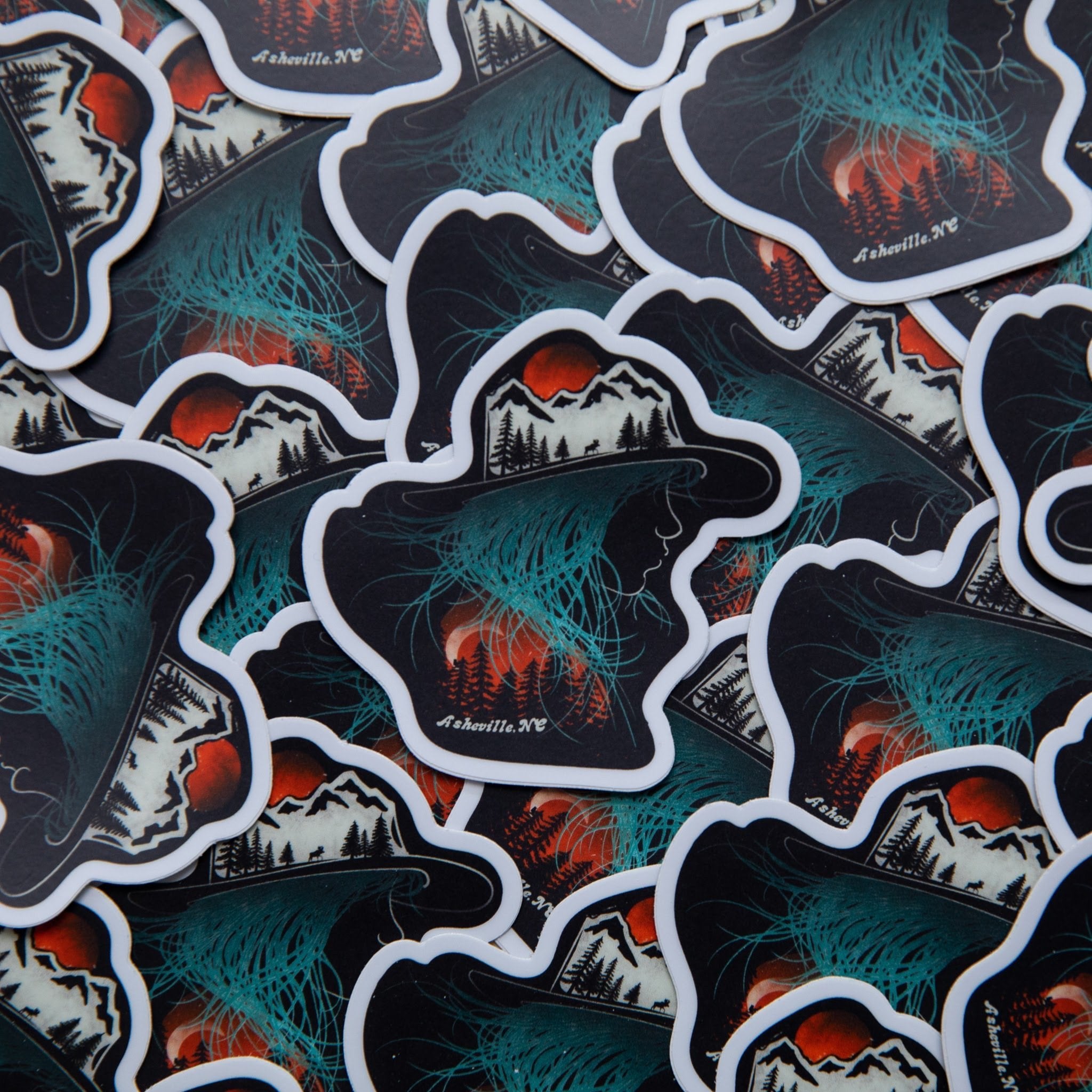 Vinyl sticker shaped like a hat with a mountain landscape and aurora-like teal glow, over a pile of identical stickers, with "Asheville NC" printed in white.
