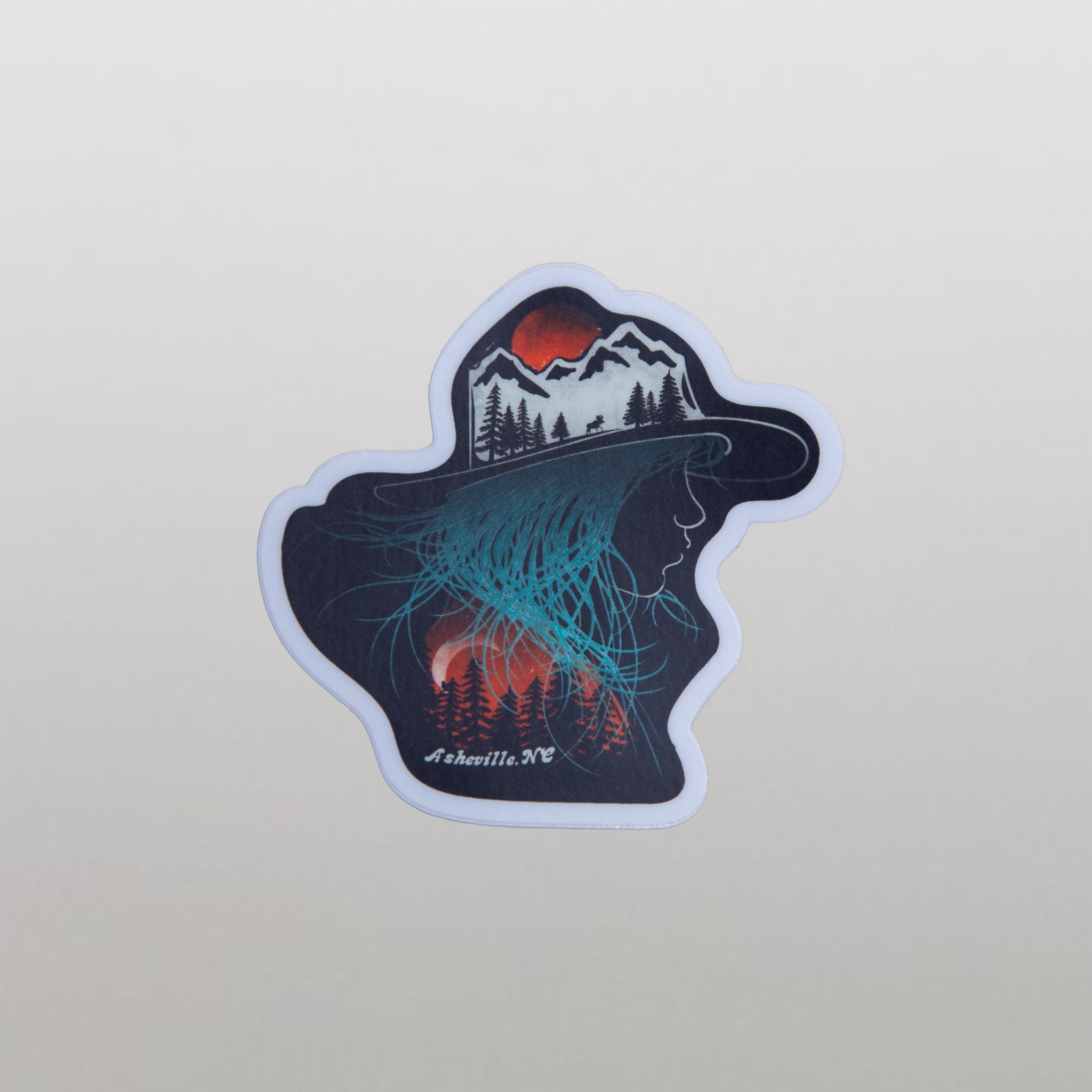 Vinyl sticker shaped like a hat with a mountain landscape and aurora-like teal glow, over a pile of identical stickers, with "Asheville NC" printed in white.