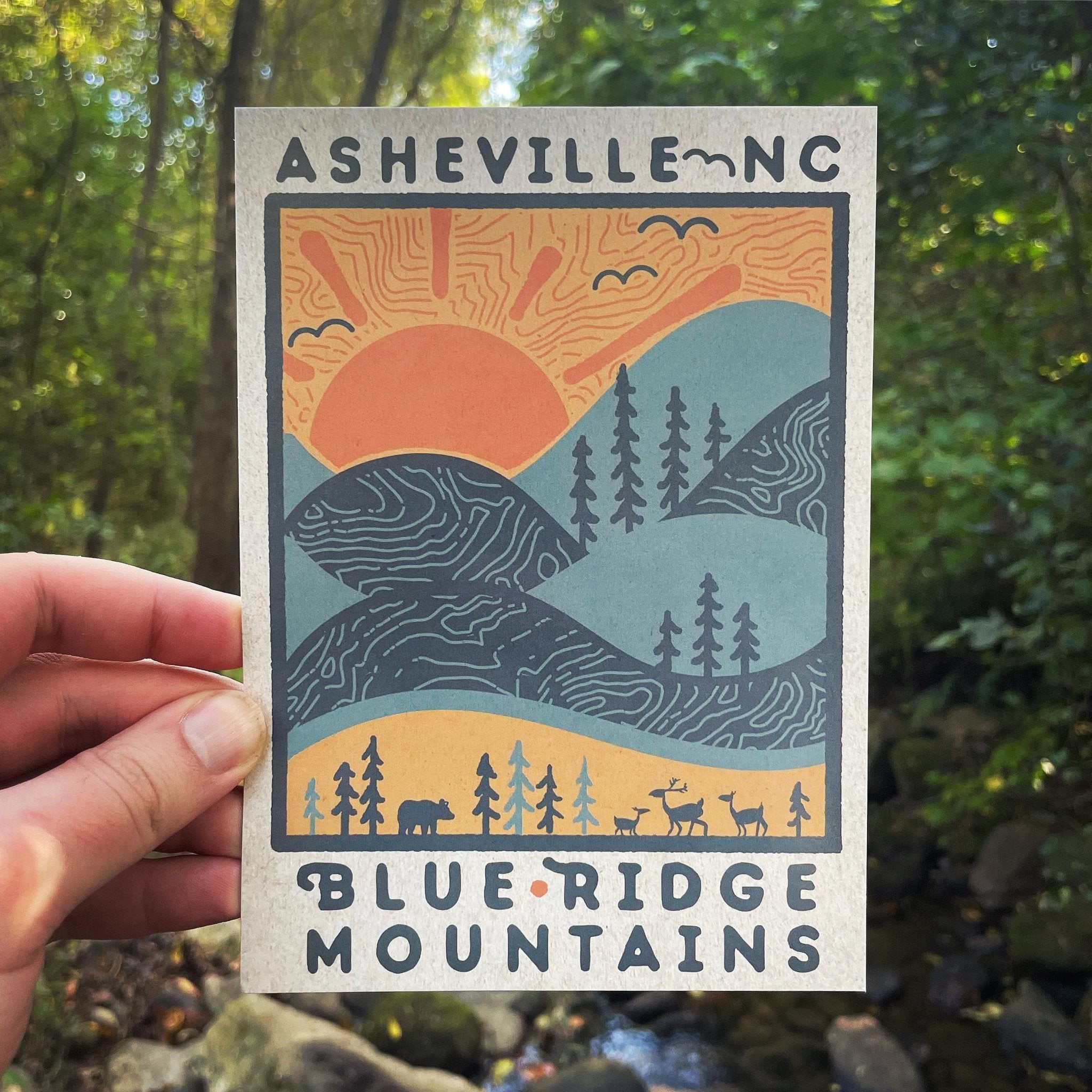 Hand holding colorful Blue Ridge Mountains postcard featuring sunrise and wildlife