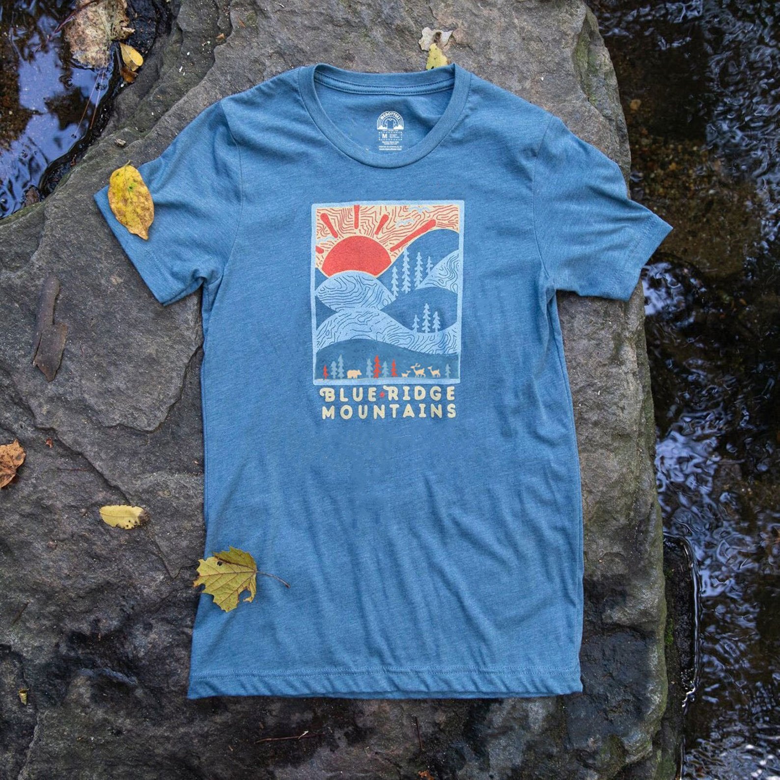 A blue crew neck t-shirt with a graphic print of the Blue Ridge Mountains and a sunset or sunrise design, laid flat on a rocky surface with a few autumn leaves around.