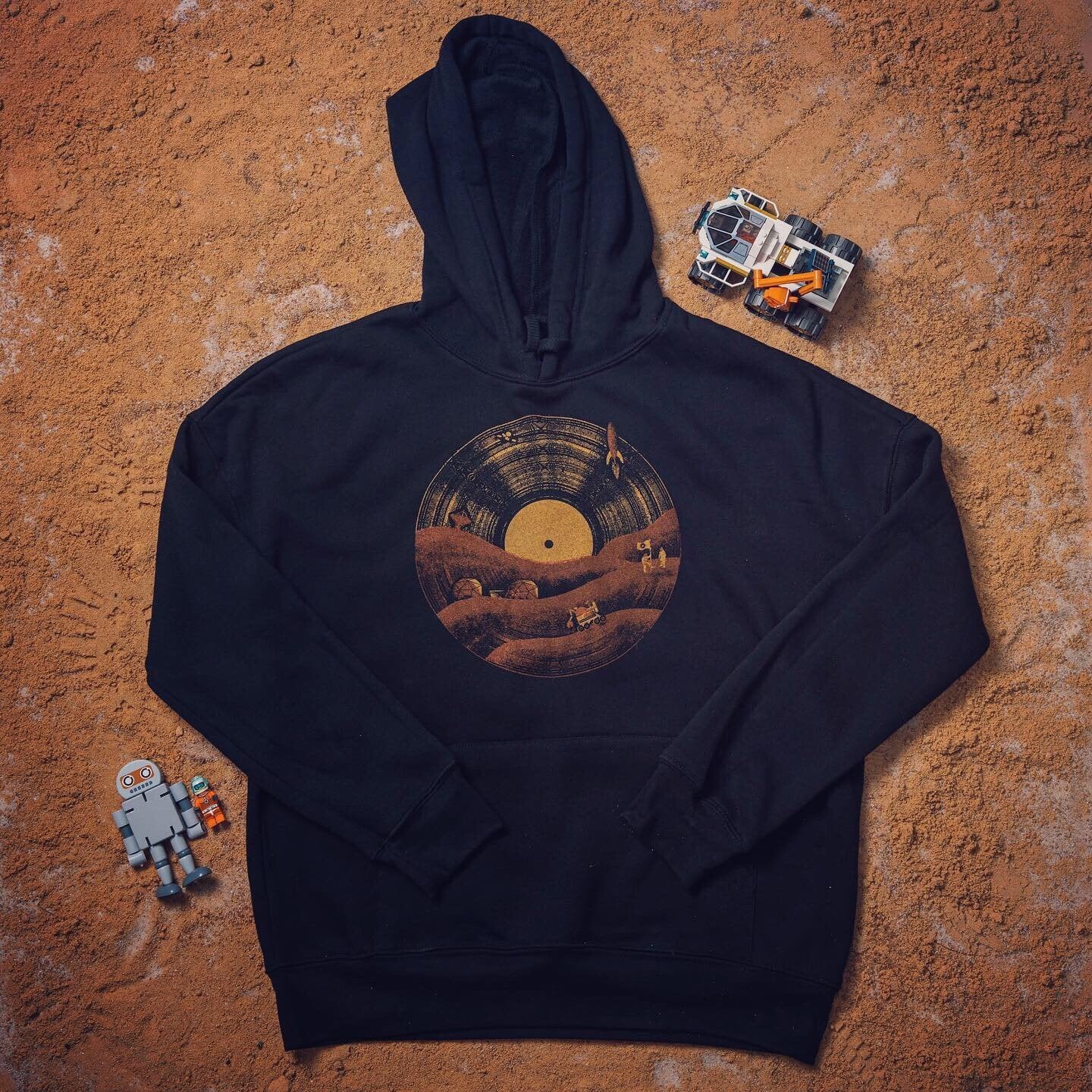 Black hoodie with a vinyl record and Mars rover illustration, laid flat on a red Martian-style surface with a robot and toy rover beside it.