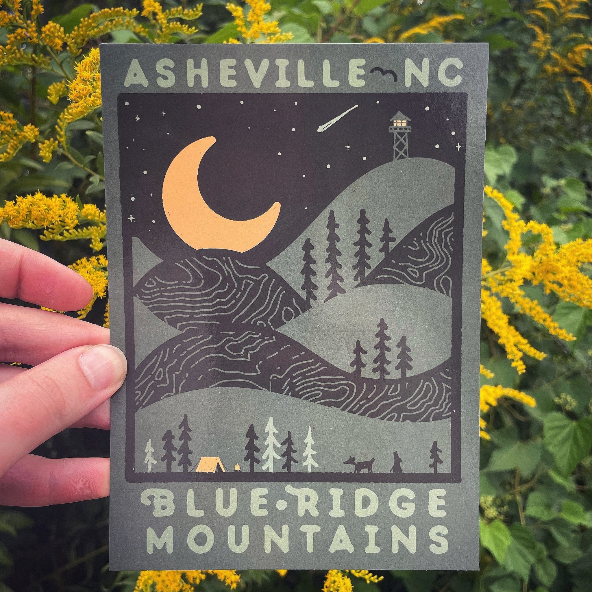Hand holding postcard with yellow crescent moon and Blue Ridge night scene, surrounded by yellow flowers