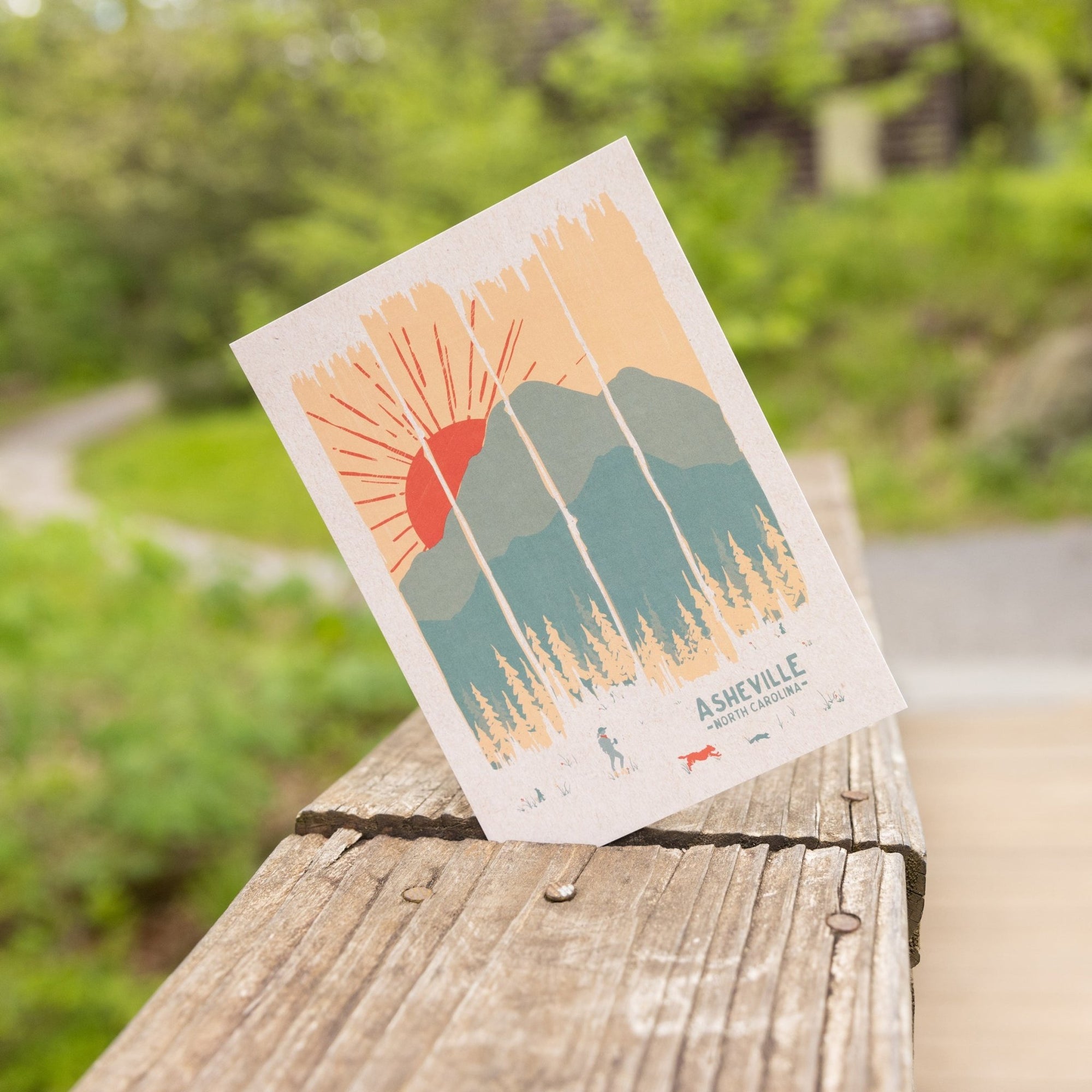 Postcard leaning against a wooden railing, featuring a stylized sun setting behind mountain peaks and pine trees, with Asheville, NC printed at the bottom.