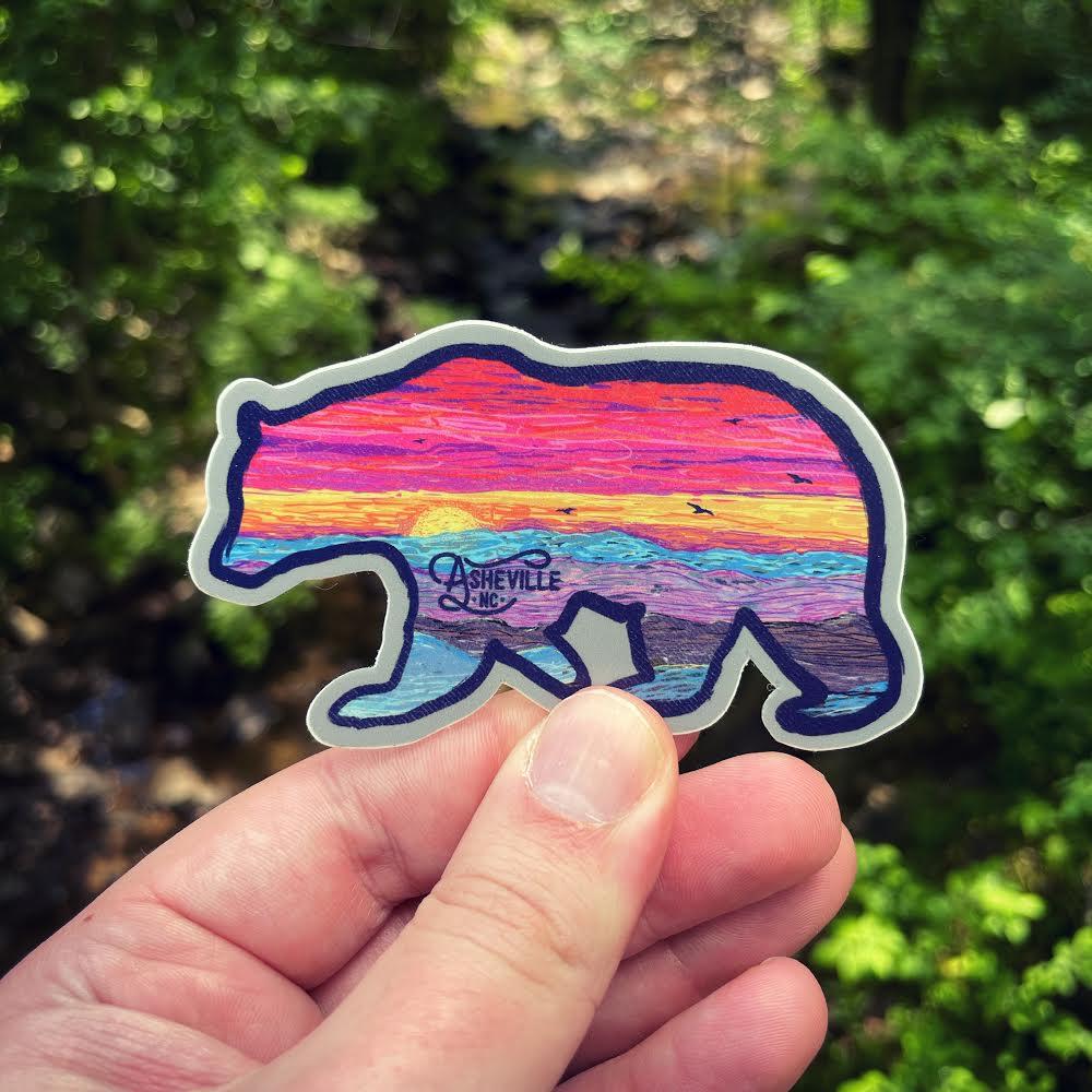 Hand holding a vibrant bear-shaped vinyl sticker with a painted sunset inside the silhouette and "Asheville NC" on the bear’s side, with a forest stream in the background.