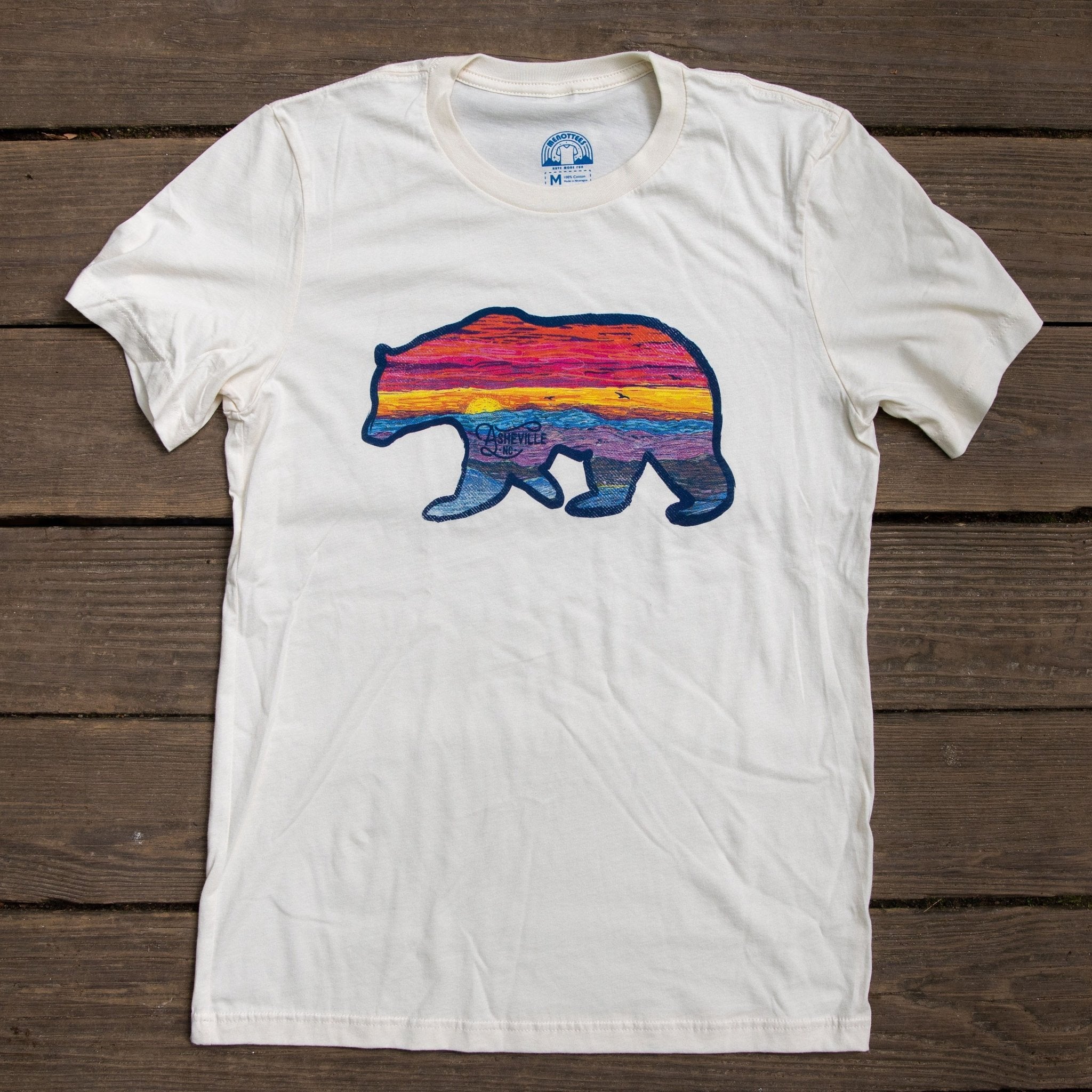 White t-shirt with colorful sunset bear silhouette laid on rustic wood”