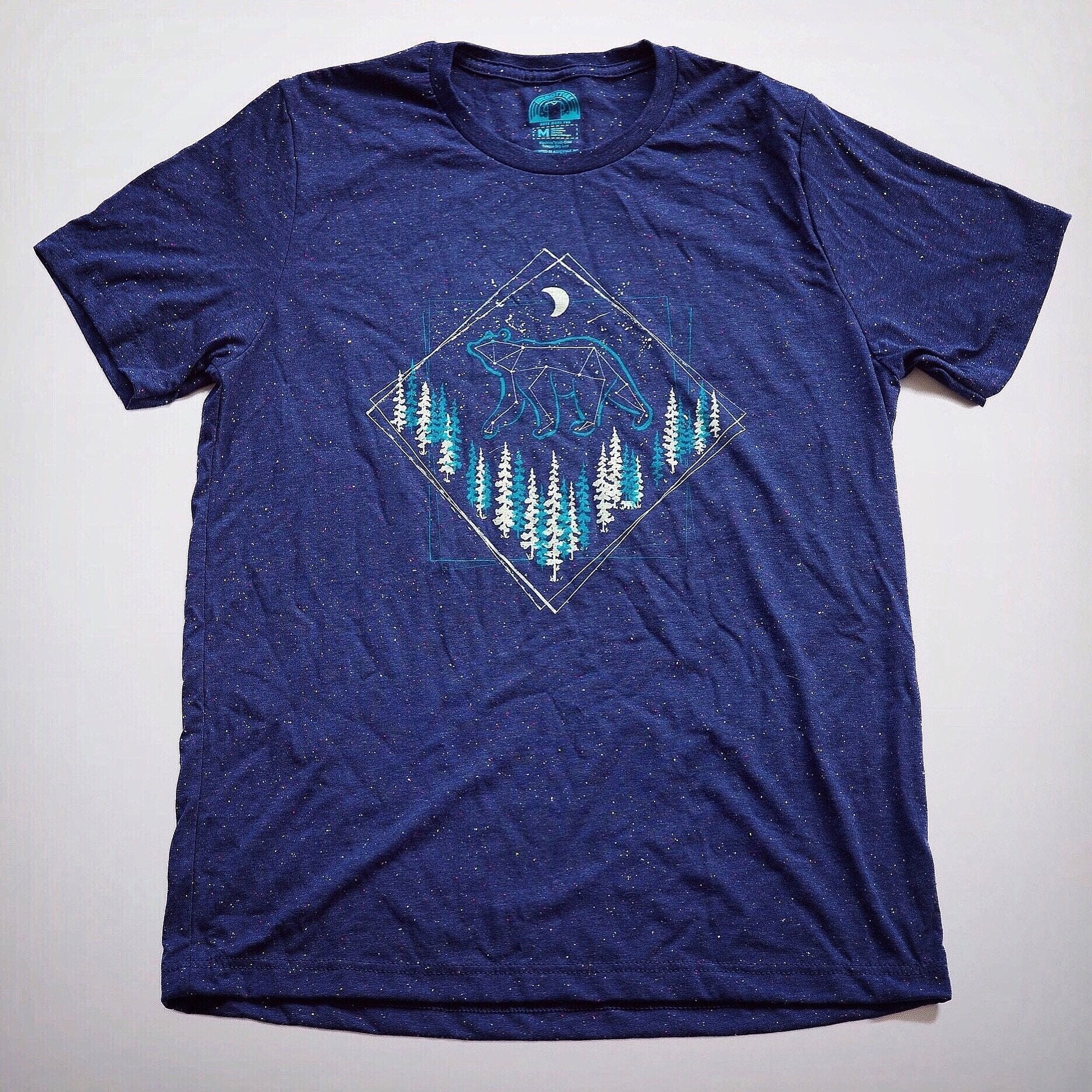 Flat-lay of navy t-shirt featuring a bear and forest graphic with moon and stars