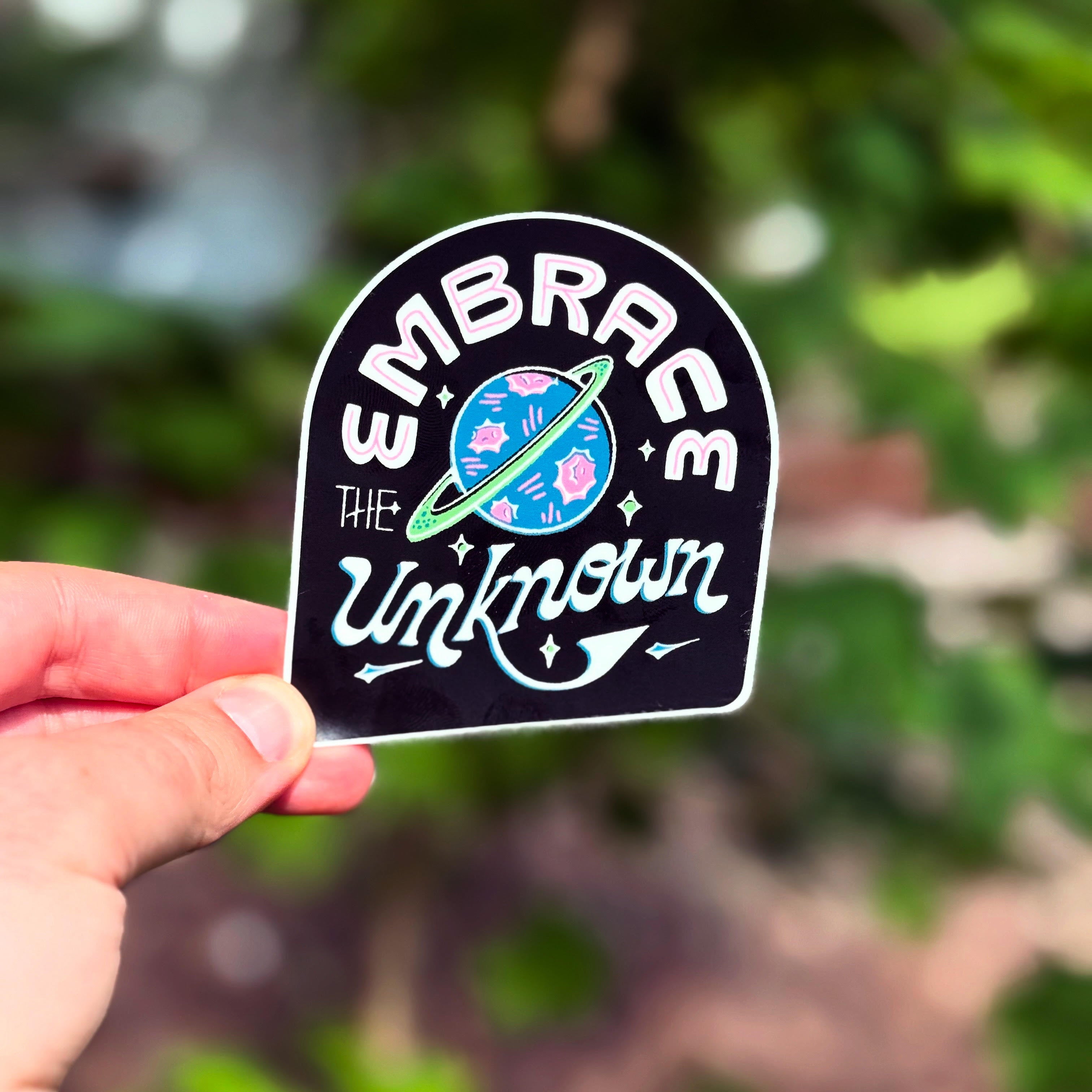 Sticker with 'Embrace the Unknown' text held by a hand against a blurred green background