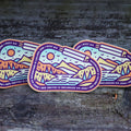 Three colorful mountain-themed stickers on a wooden surface