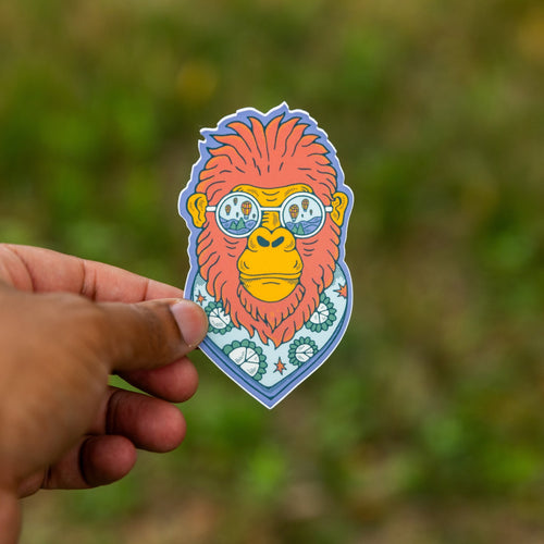 Close-up of a hand holding a vinyl sticker of a red-orange Bigfoot wearing balloon sunglasses and a patterned scarf, with a blurred green background.


