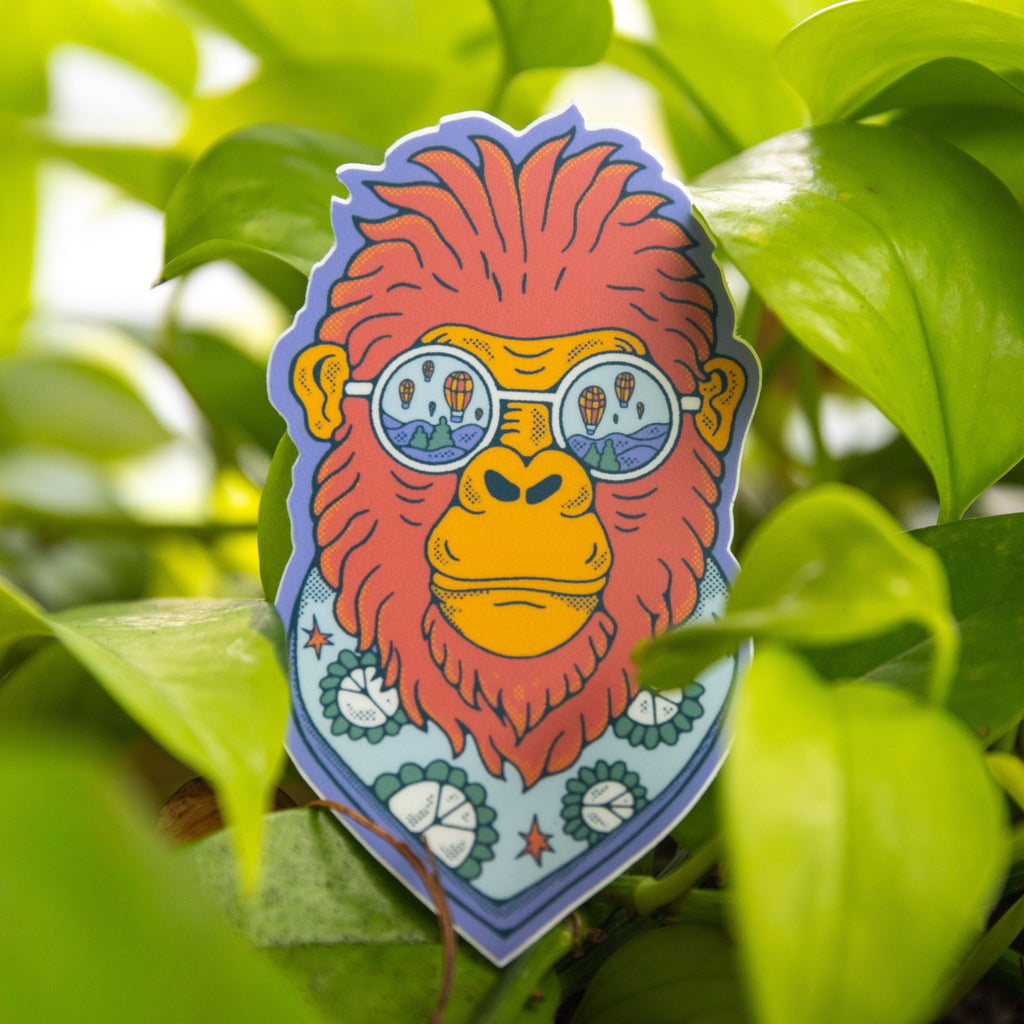 Sticker of Bigfoot with balloon sunglasses and a scarf, placed in direct sunlight against green foliage.

