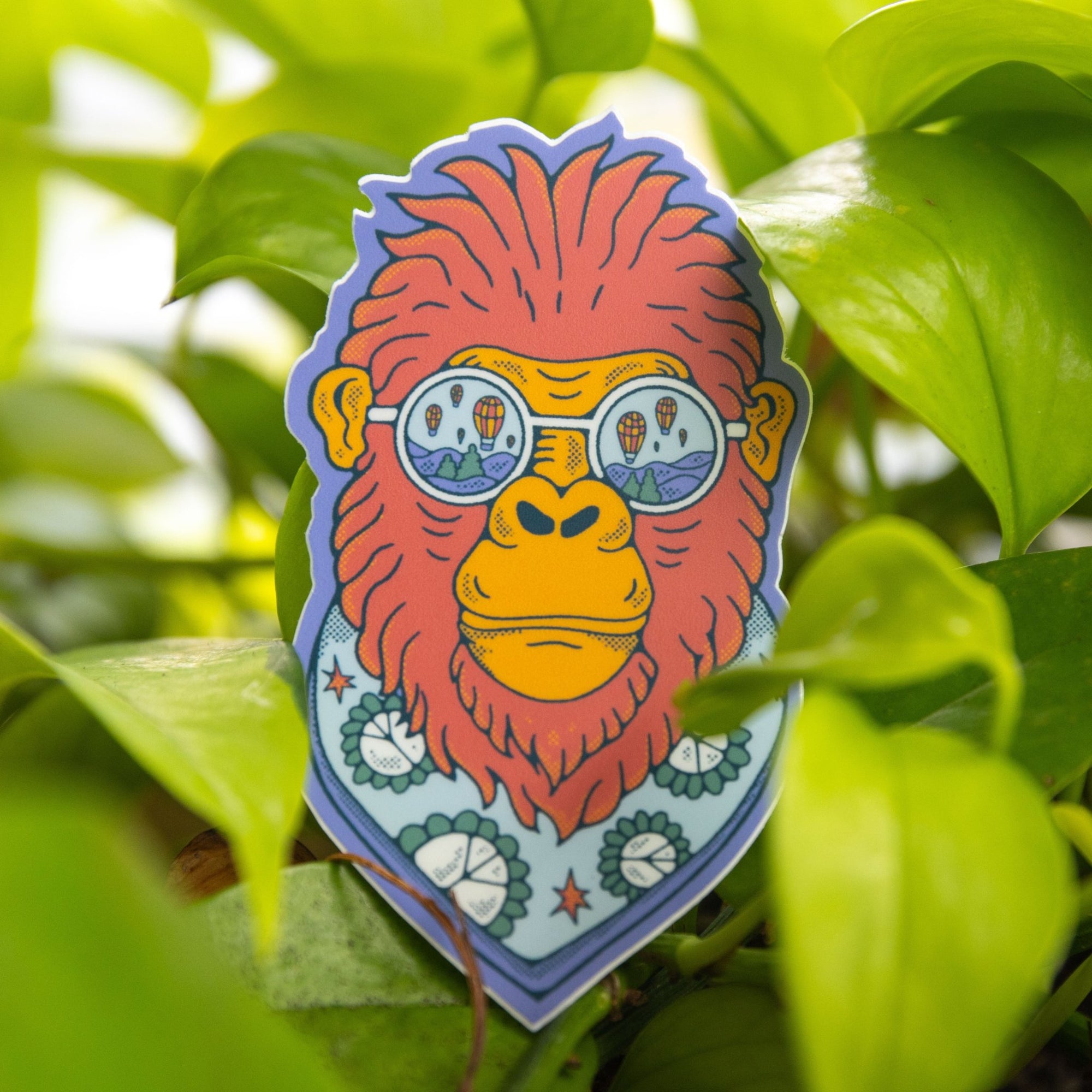 Sticker of Bigfoot with balloon sunglasses and a scarf, placed in direct sunlight against green foliage.

