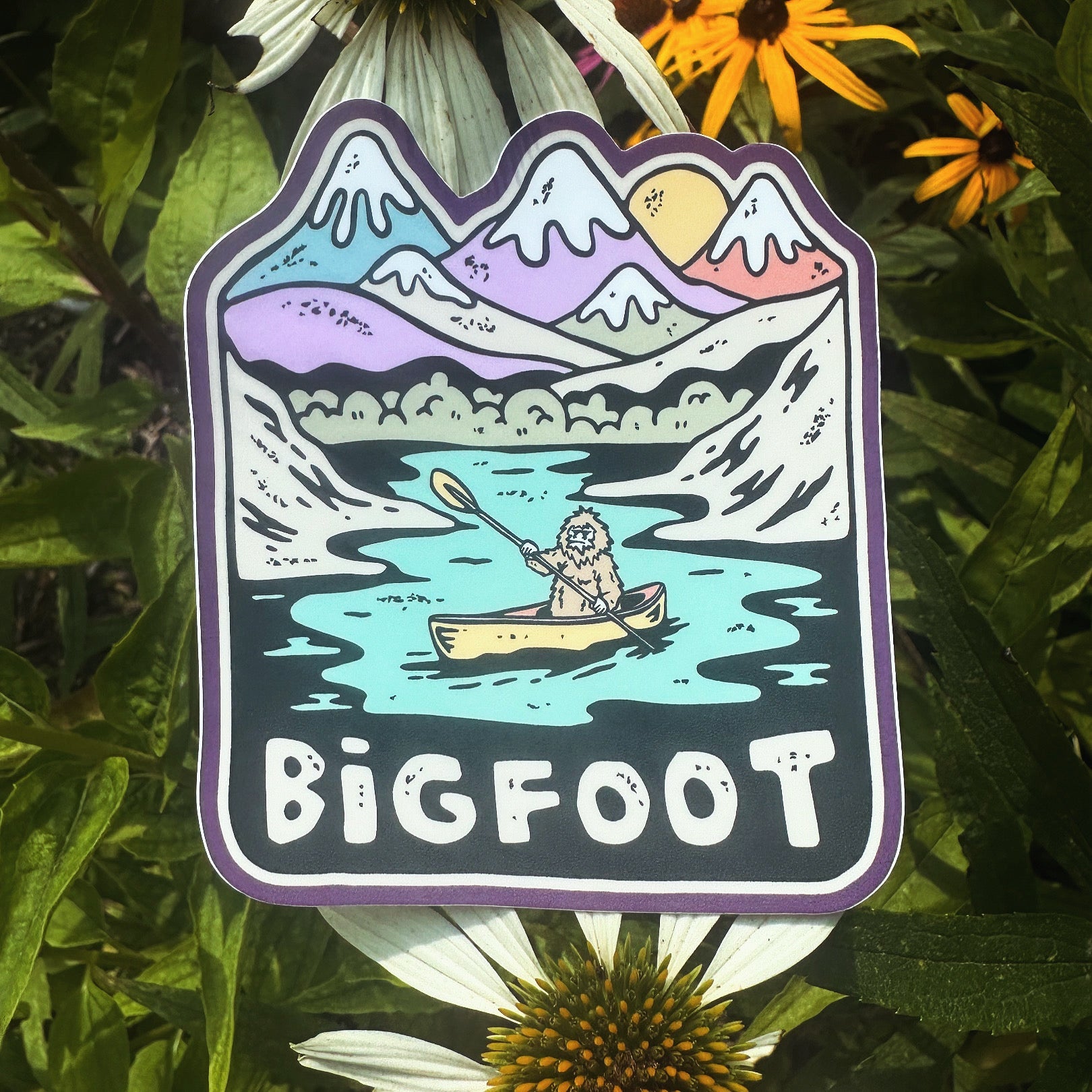 Bigfoot Canoe Trip sticker propped in flowers, blending with natural surroundings.

