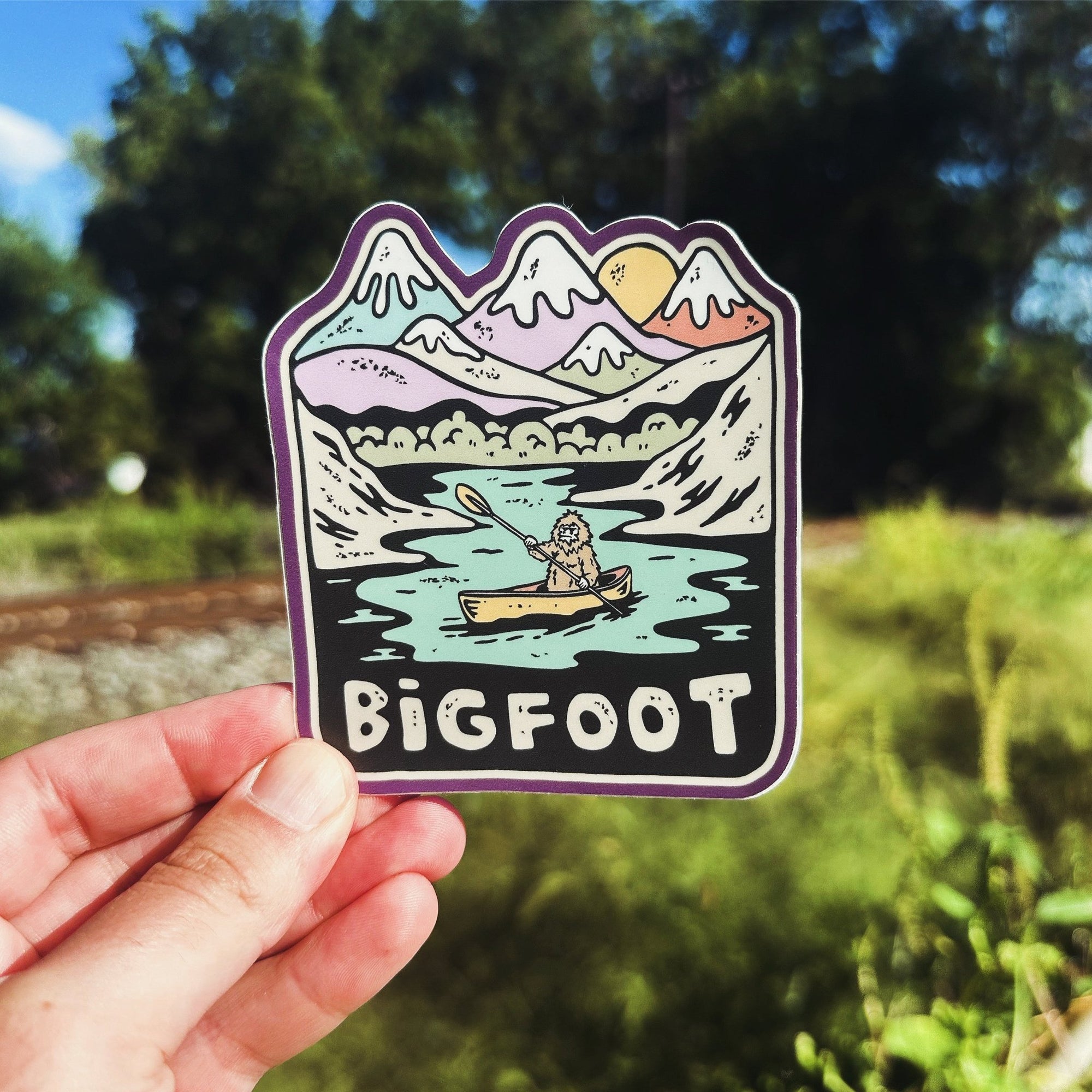 A hand holding a vinyl sticker with an illustration of Bigfoot and a canoe on a sticker, with mountains and a sun in the background.