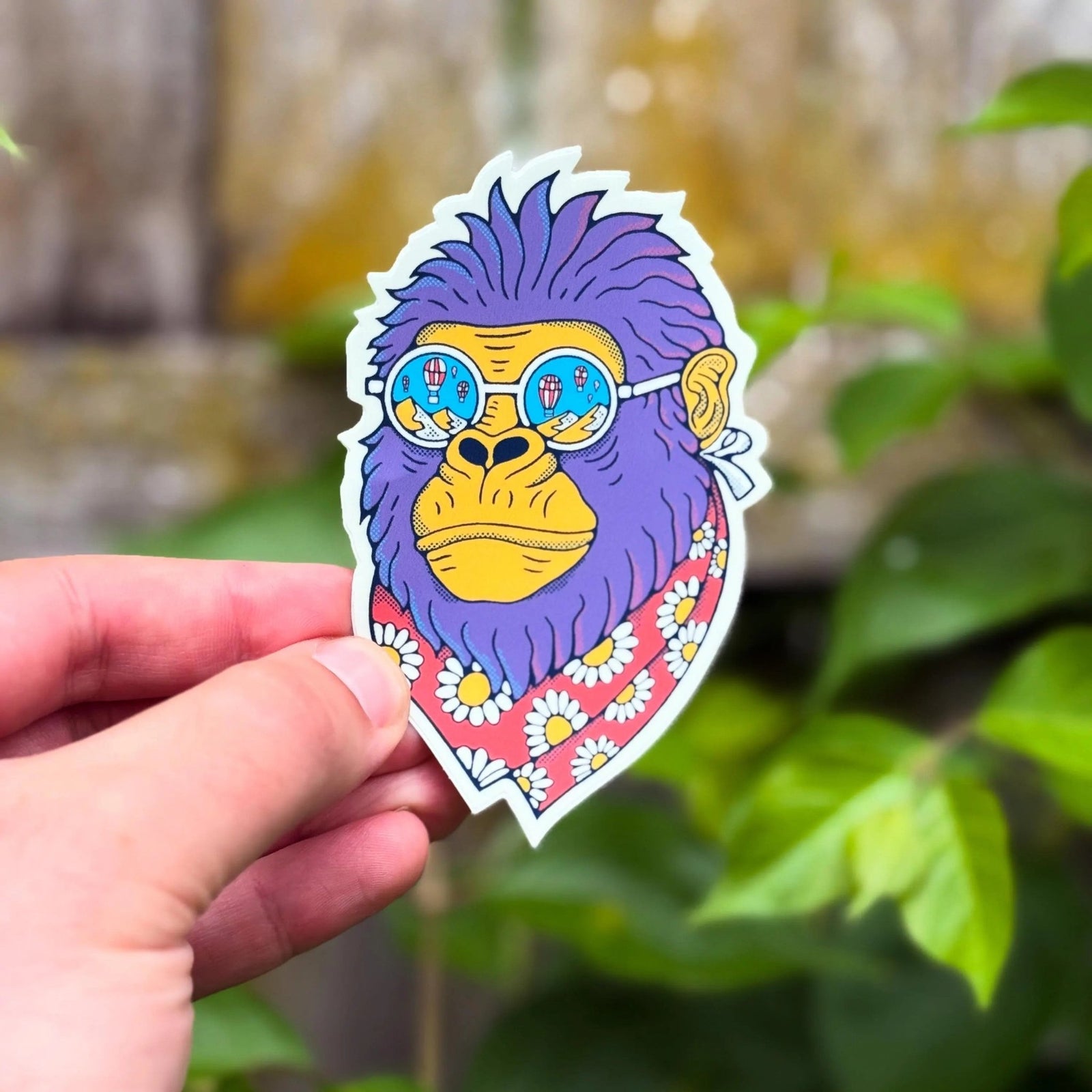 Hand holding a colorful sticker of a monkey with sunglasses against a natural background