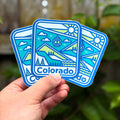 Three Colorado-themed stickers held by a hand with a blurred natural background