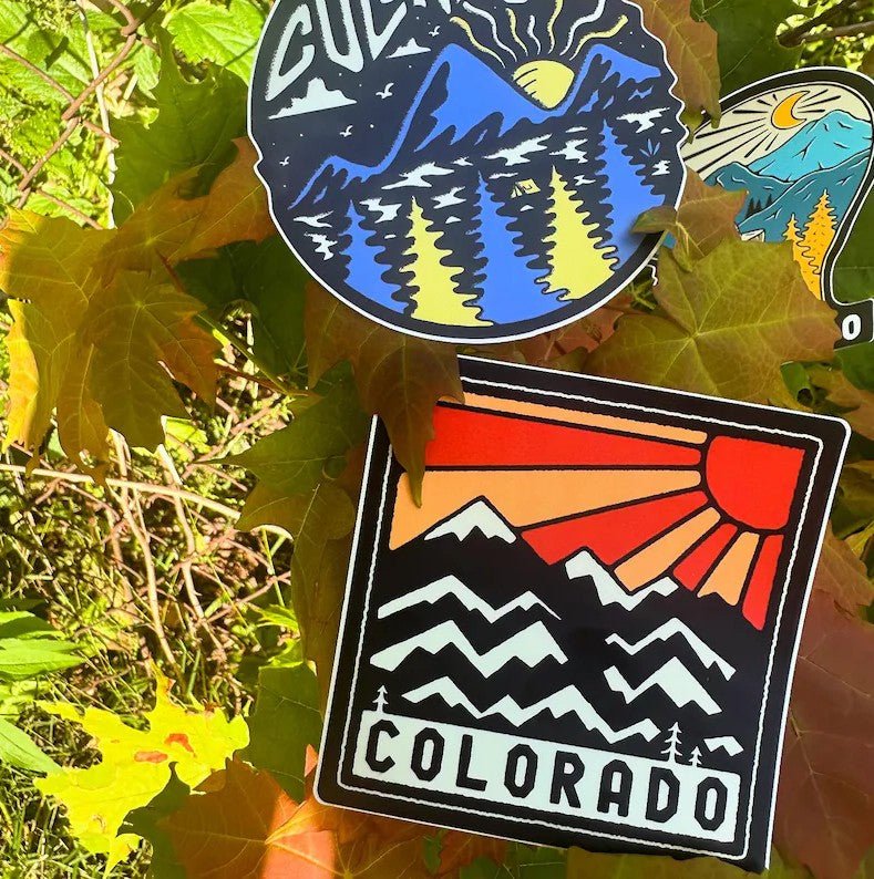 Two stickers with mountain and sun designs on a leafy background