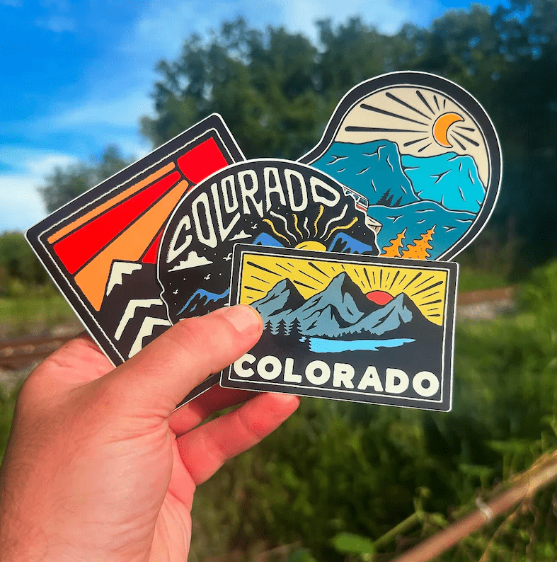 Hand holding Colorado-themed stickers with mountain and sun design against a natural background.
