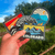 Hand holding Colorado-themed stickers with mountain and sun design against a natural background.
