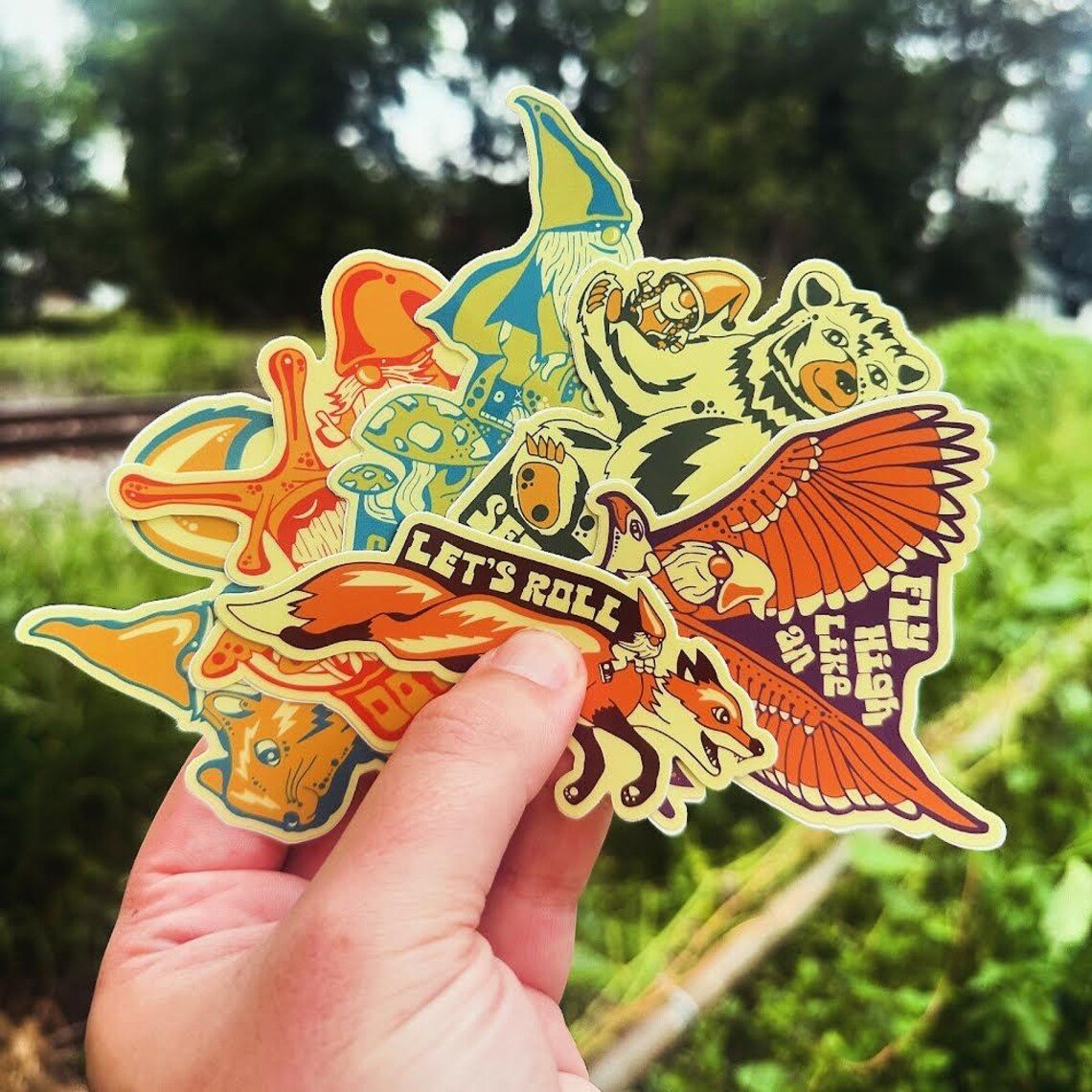 Hand holding full set of 6 colorful vinyl gnome stickers in front of train tracks