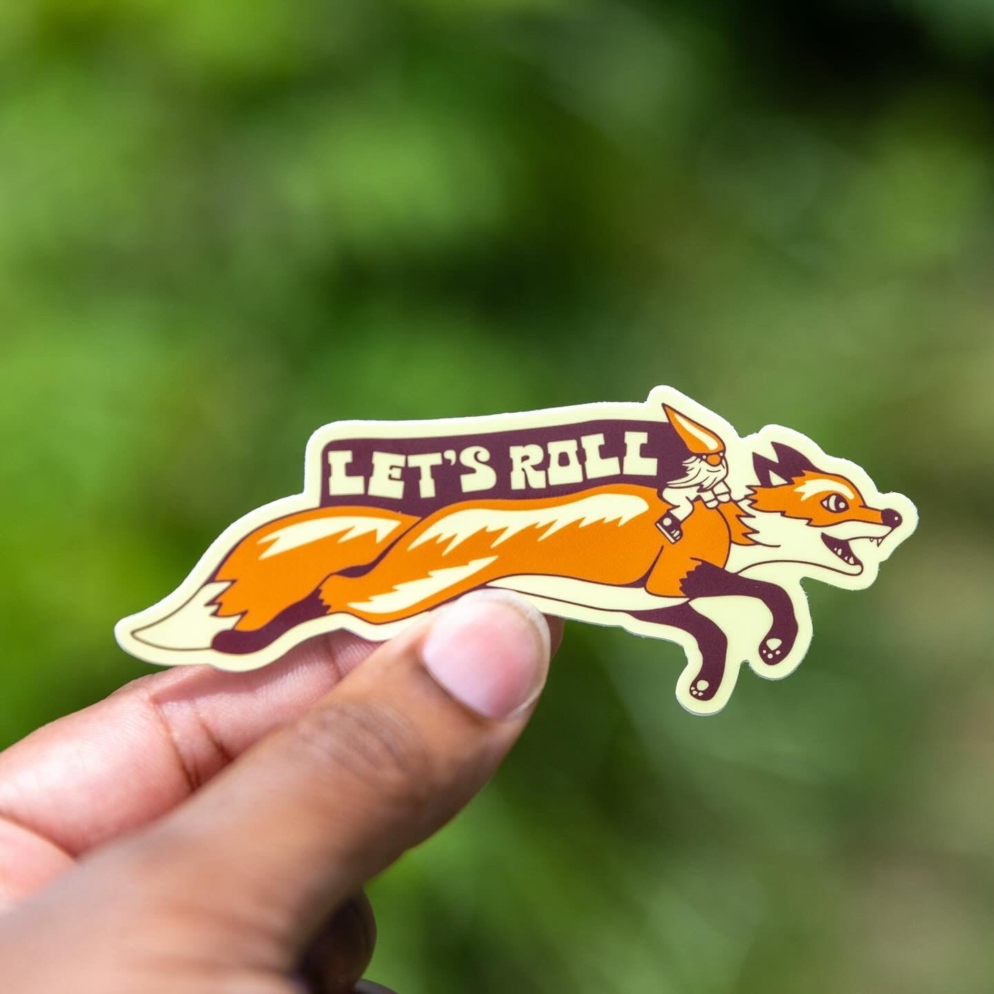 Hand holding vinyl sticker featuring a gnome riding a running fox with “Let’s Roll” text