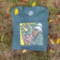 Folded green New Beginnings shirt with colorful sunrise and flower design on grass