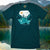 Teal t-shirt with nature graphic design in front of a lake and mountains