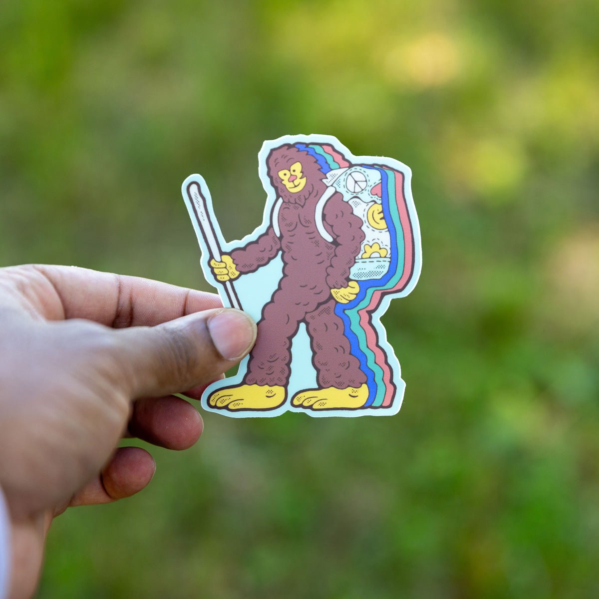 A sticker depicting a cartoonish bigfoot character with a colorful backpack, being held by a hand against a blurred natural background.