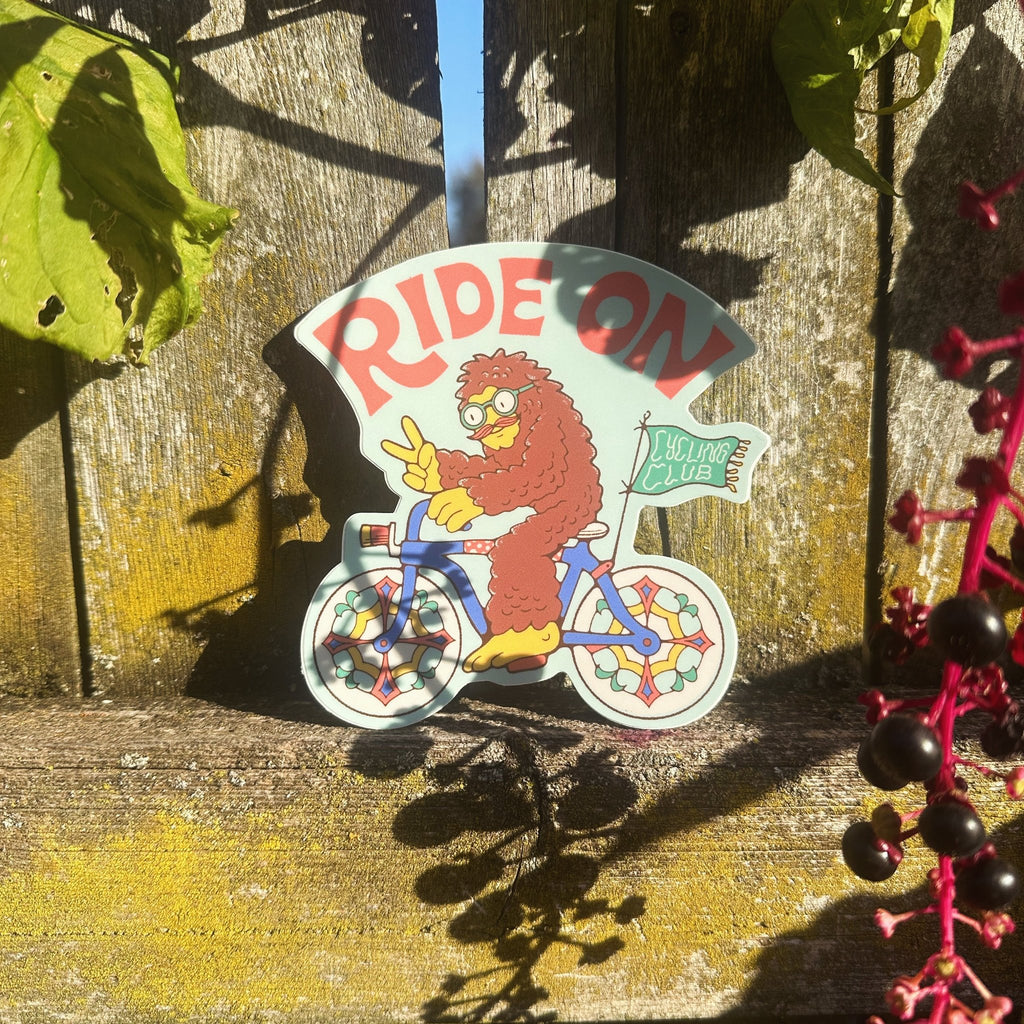 Bigfoot riding a bike sticker propped up outdoors with a wooden fence and sunlight behind.

