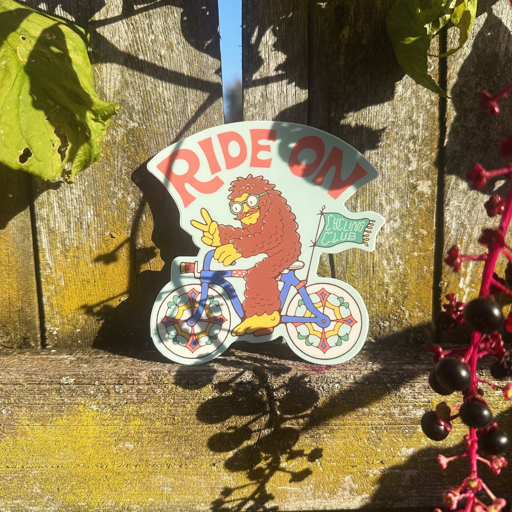 Bigfoot riding a bike sticker propped up outdoors with a wooden fence and sunlight behind.

