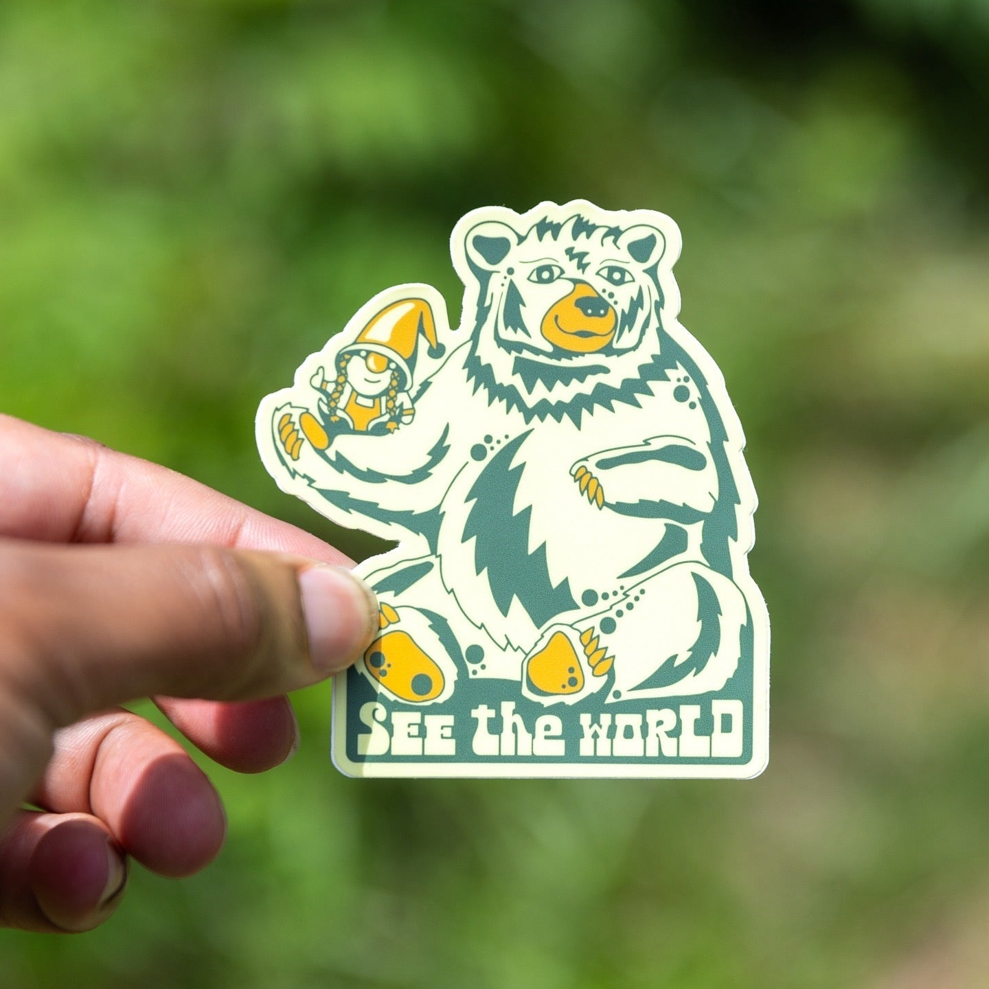 A hand holding a vinyl sticker with a gnome and a bear design, with the text 'See the World'.